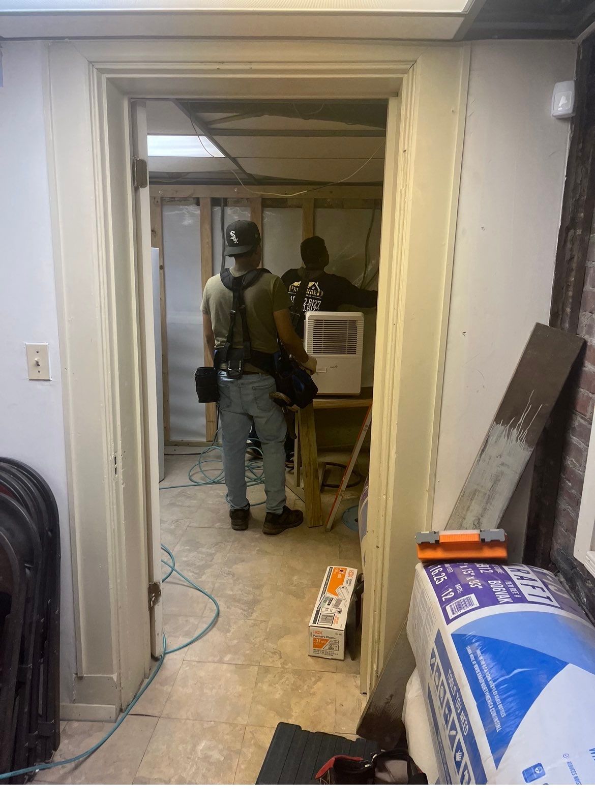 Two construction workers inside a room under renovation; visible studs, insulation, and tools.