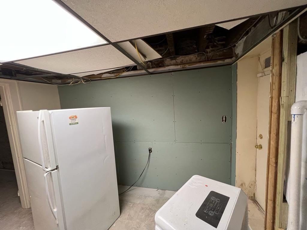 Basement room with a refrigerator, damaged ceiling, and exposed wall studs.