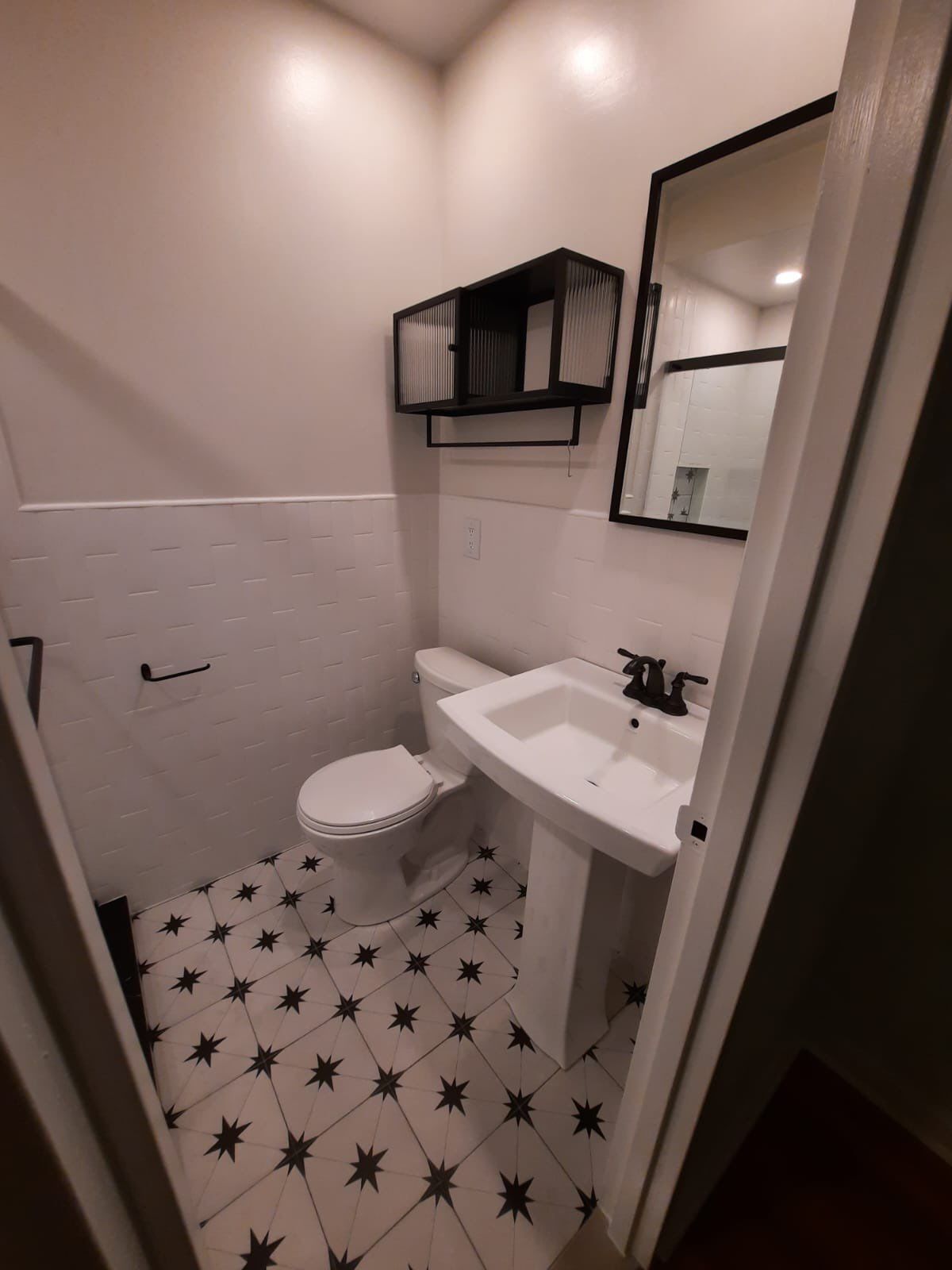 Small bathroom with black and white star-patterned tile floor, pedestal sink, toilet, and black-framed mirror.