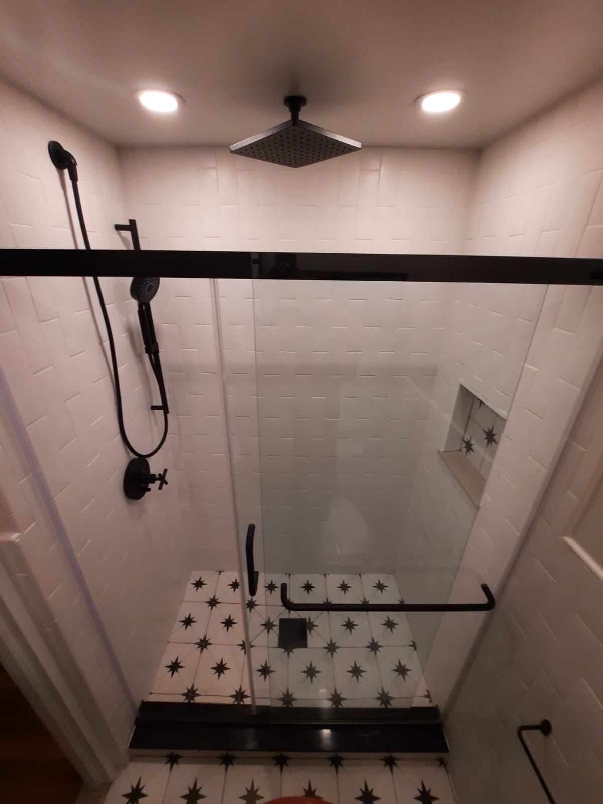 Modern shower stall with black fixtures, glass door, and white tiled walls. Black and white patterned floor.