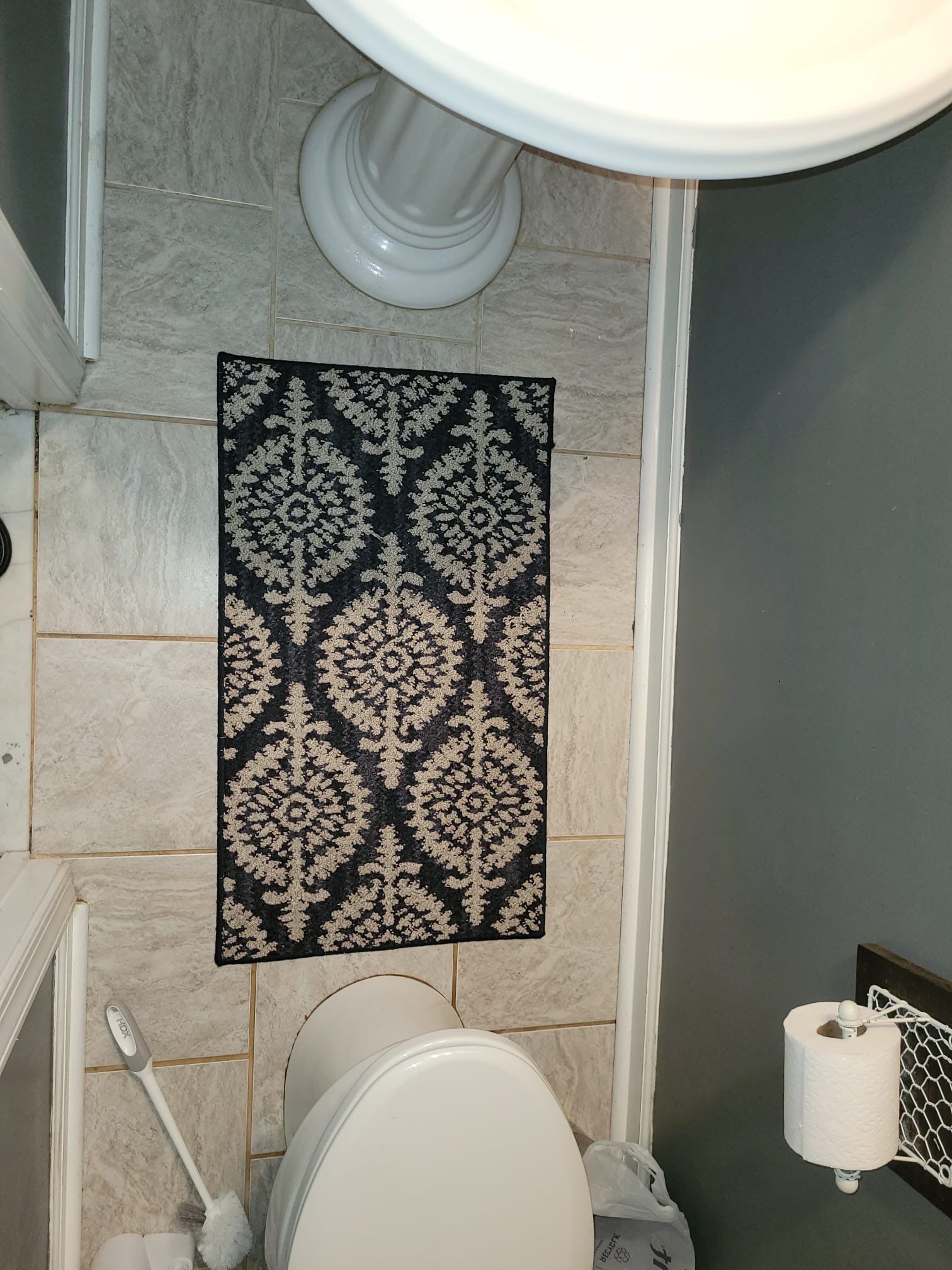 Small bathroom with toilet and decorative rug. Tiles and a roll of toilet paper are visible.