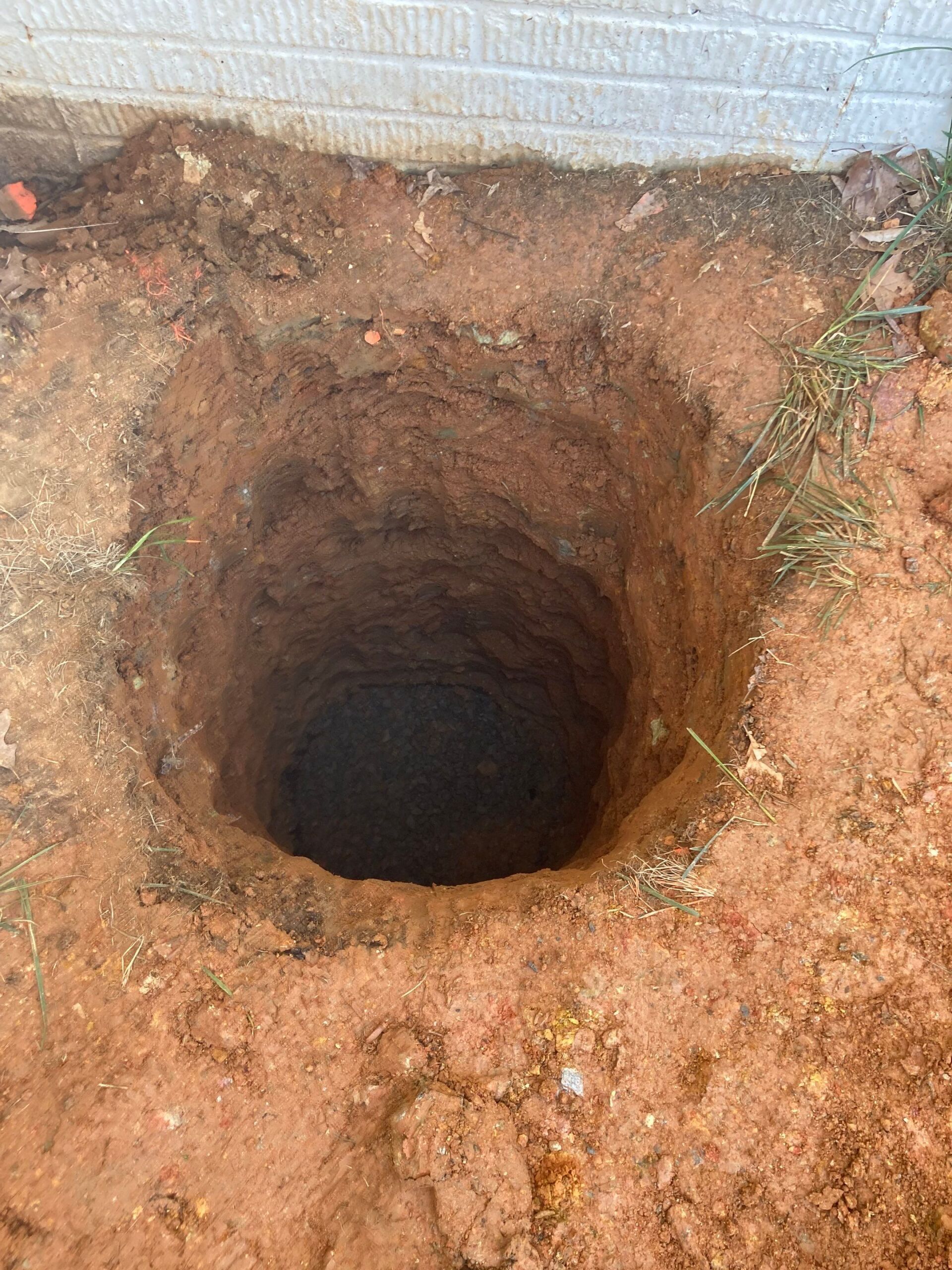 A deep, square hole dug in reddish-brown soil next to a light-colored brick wall.