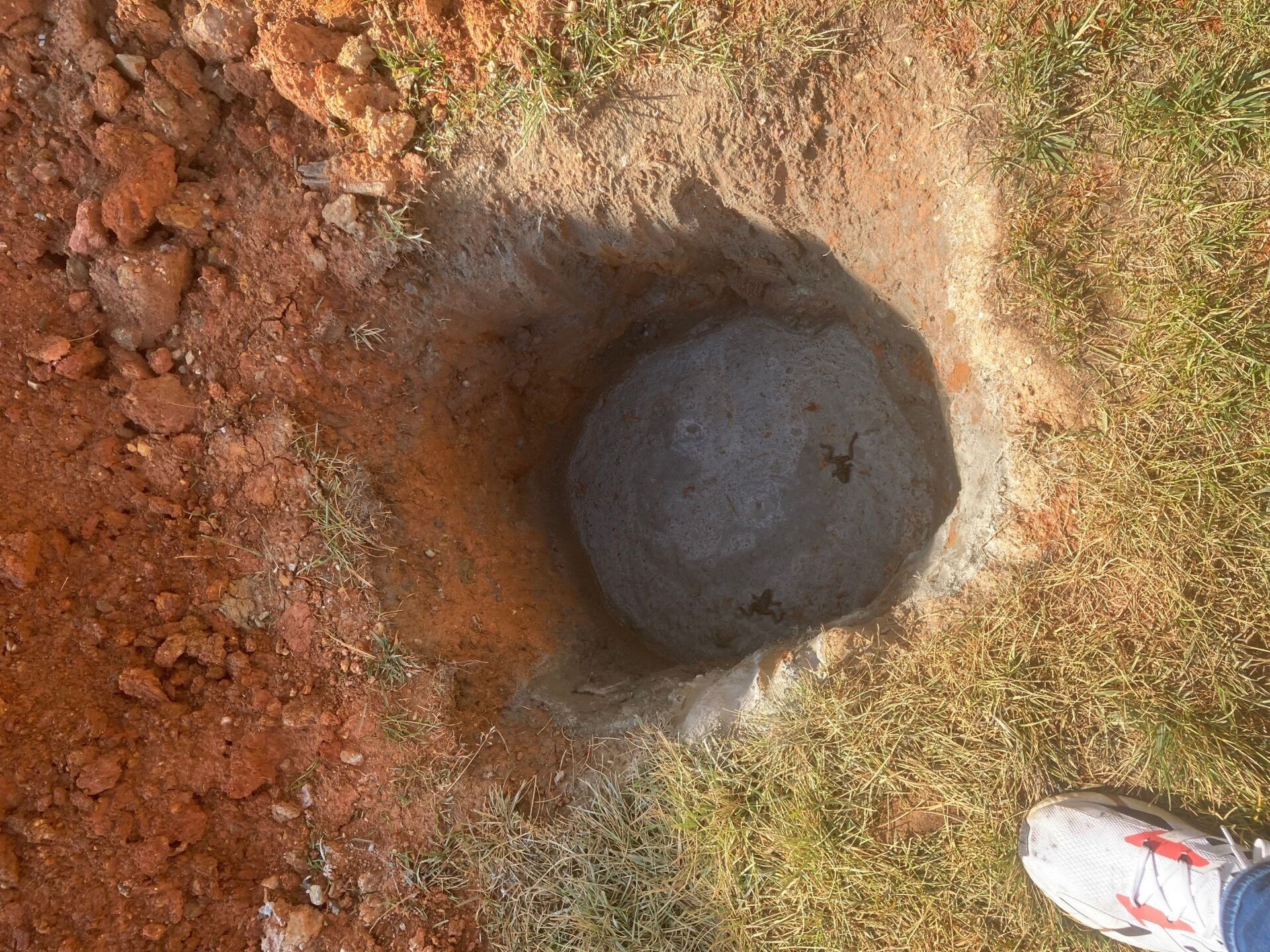 Hole in the ground with a large, round gray object inside, likely concrete, in a grassy area.