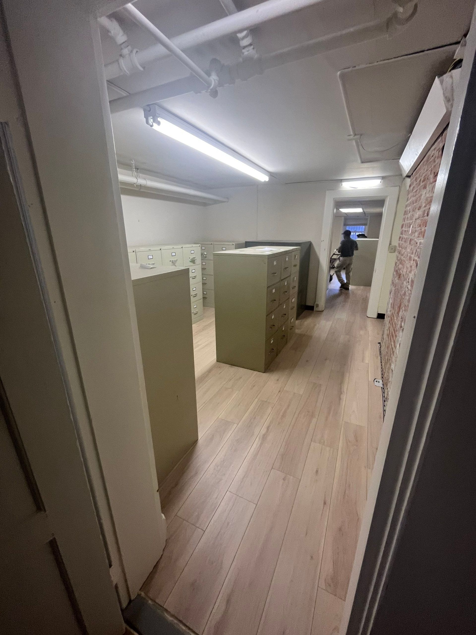 Long, narrow room with filing cabinets, a person in the distance, and light-colored wood-look flooring.
