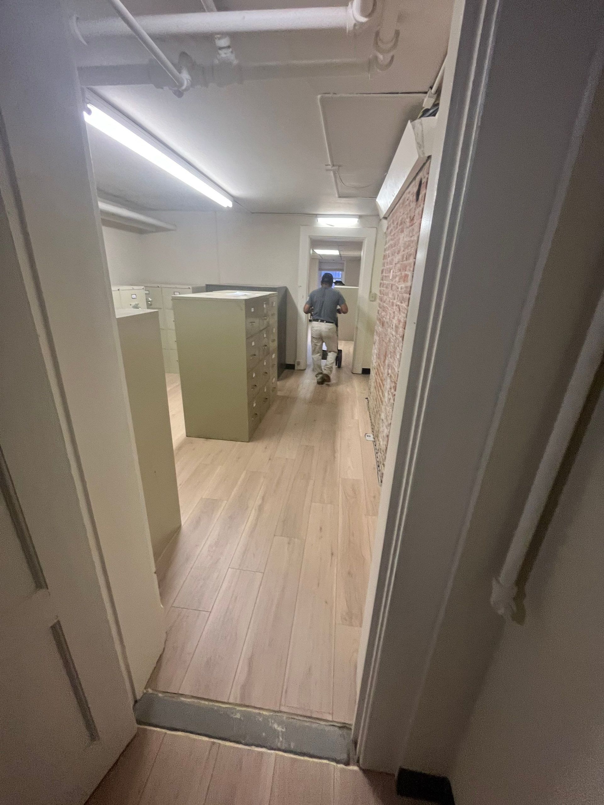 A person walks down a long hallway with light wood floors. File cabinets line the left side.