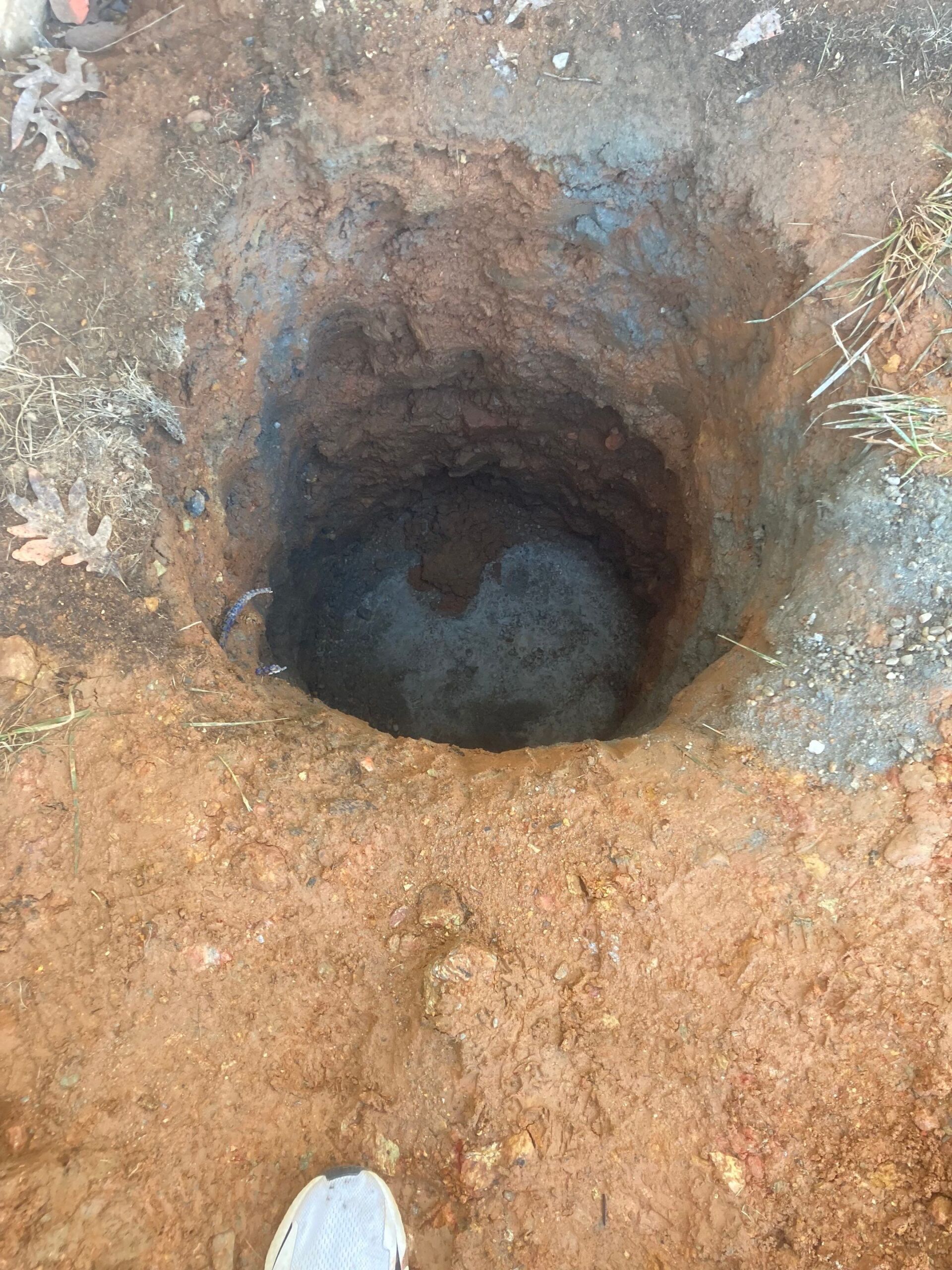 A deep, round hole in reddish-brown earth, with a dark interior and loose soil around the edges.