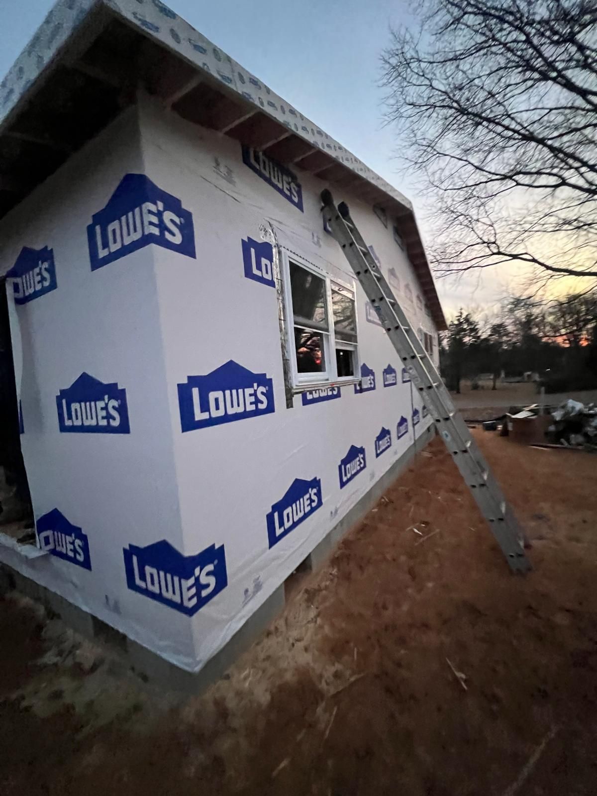 Construction site: building wrapped in Lowe's branded weather barrier; ladder propped against wall; dusk setting.