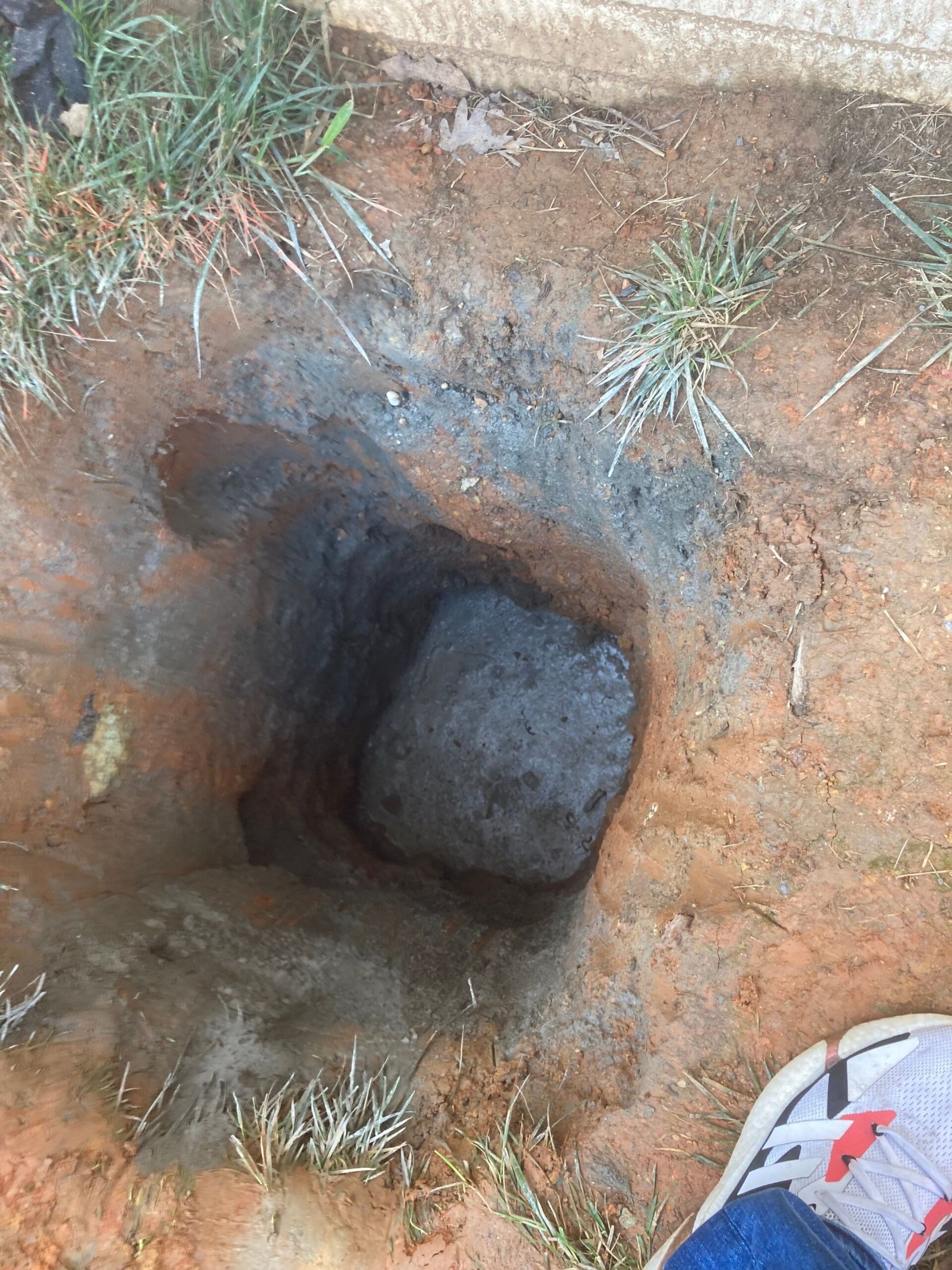Muddy hole in the ground, dark center, surrounded by reddish dirt and grass, foot in frame.