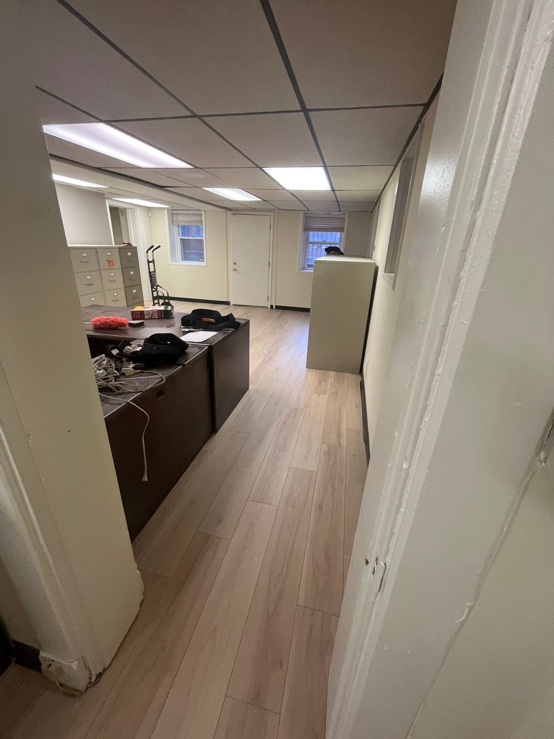A long, narrow office hallway with wood floors, white walls, and a dropped ceiling with fluorescent lights.