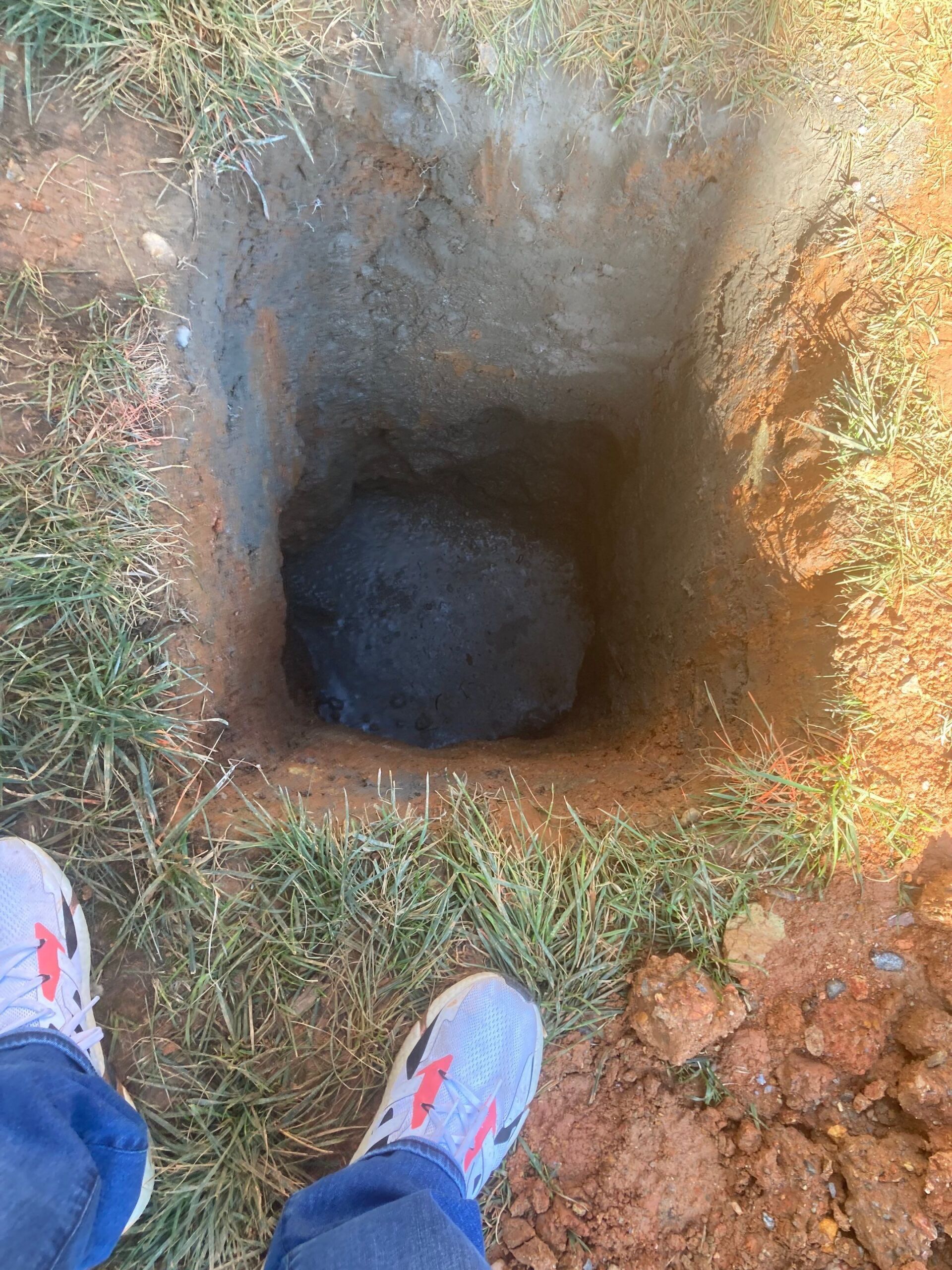 Hole in the ground with a large, dark stone at the bottom. Person's feet are visible on the grass.