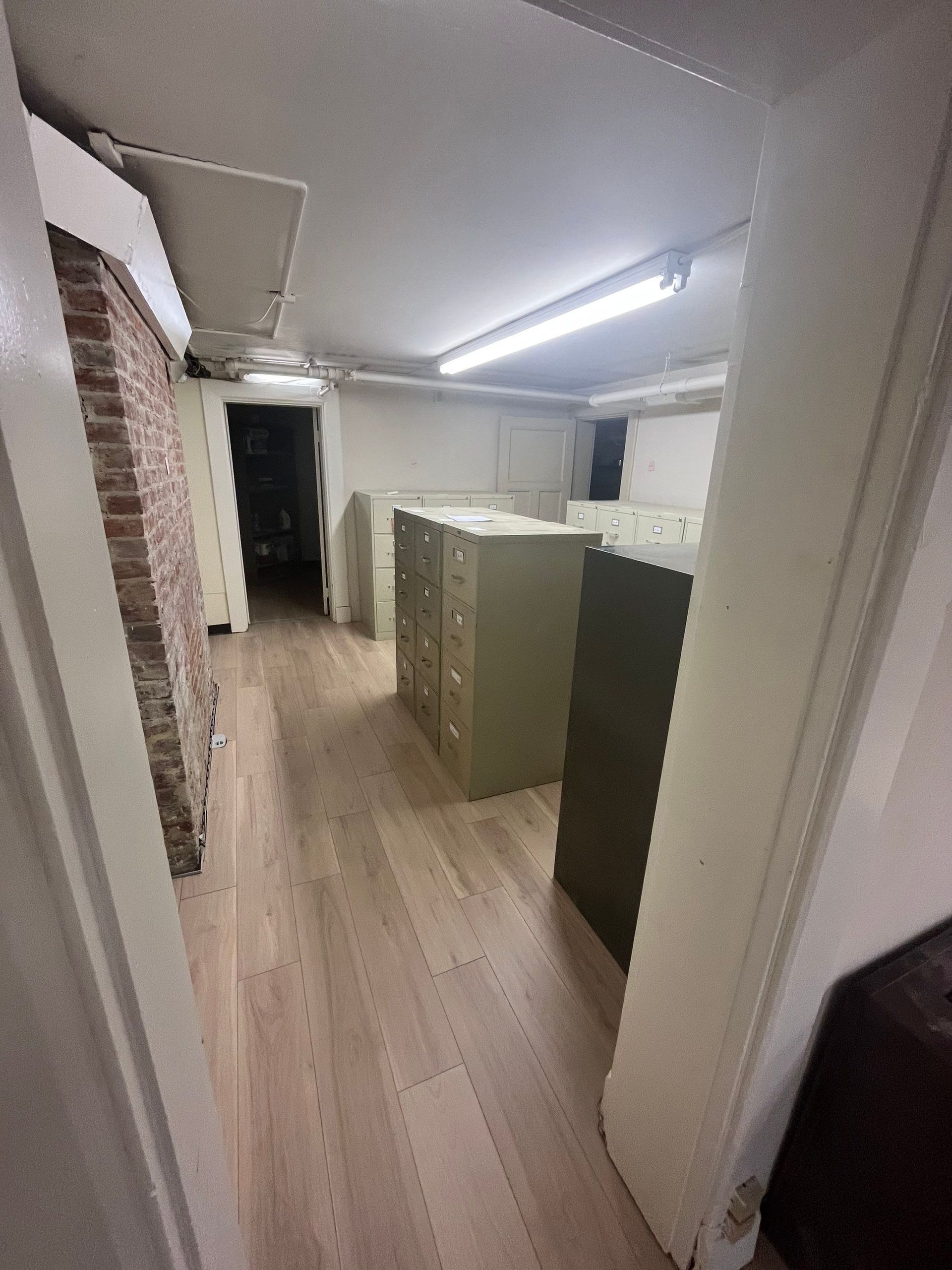 Long, narrow office with light wood floors, file cabinets, and exposed brick wall.