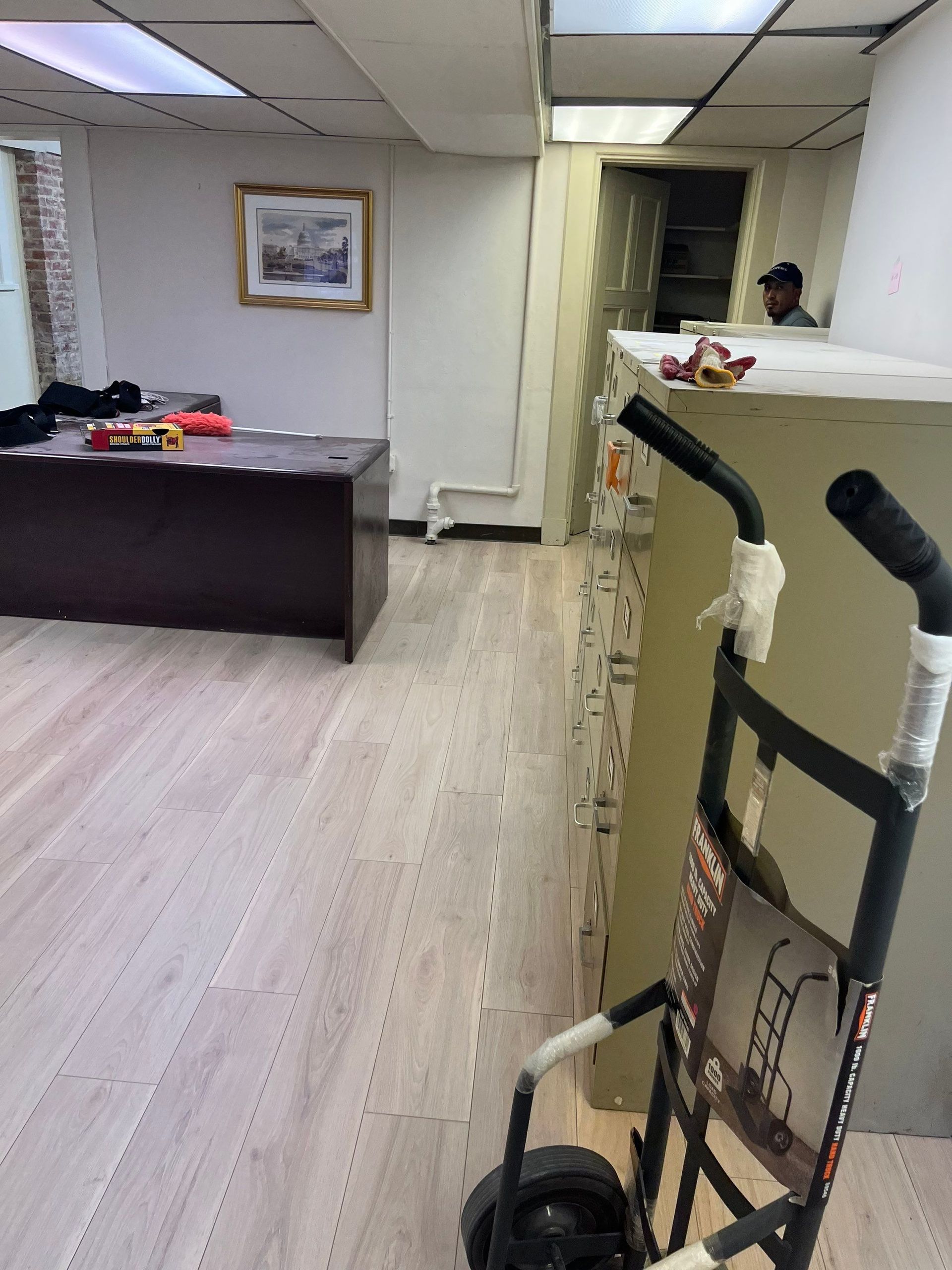 Office interior with a desk, cart, and person visible near a counter. Light-colored flooring.