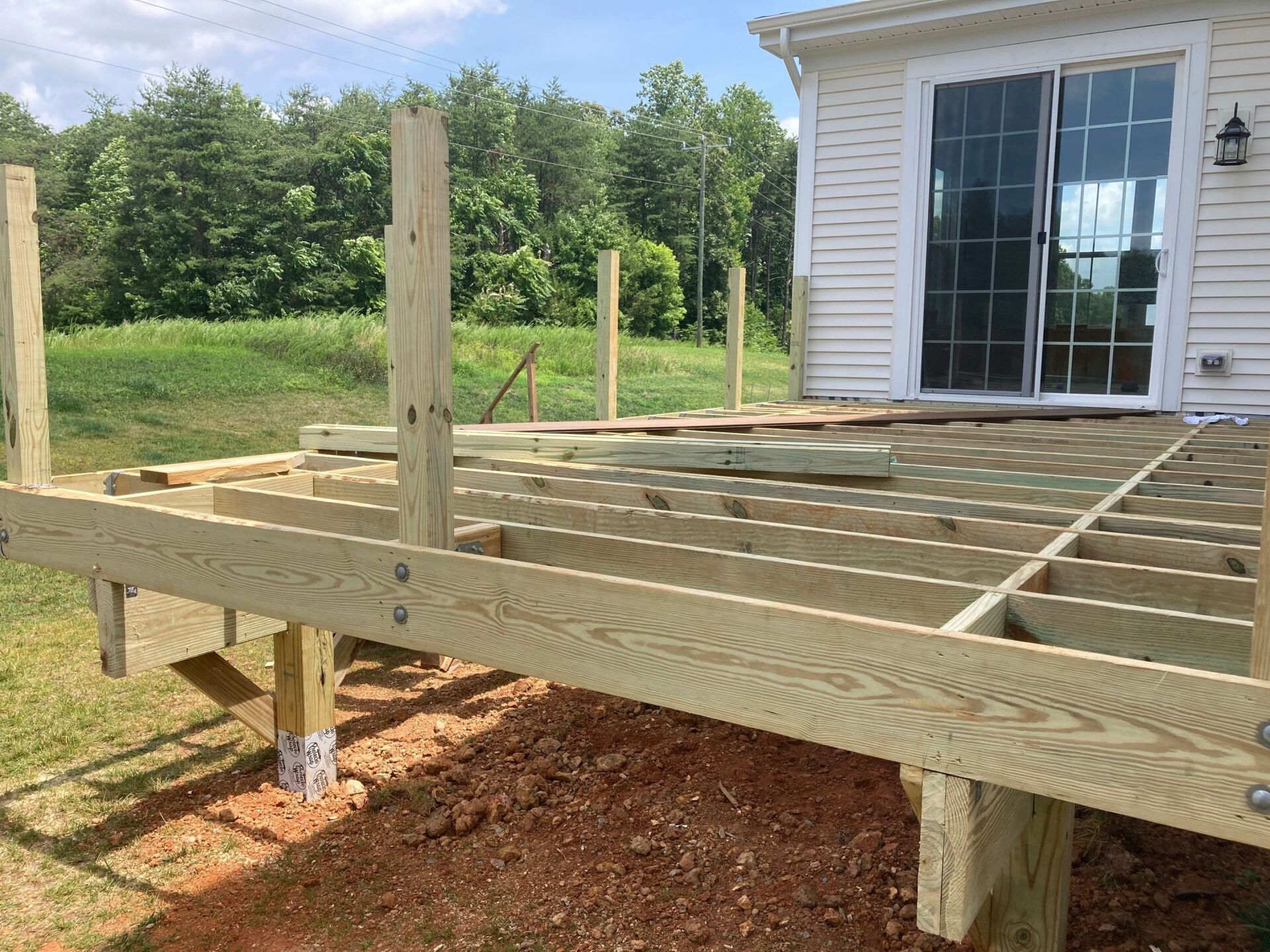 Wooden deck under construction next to a house with a sliding glass door; sunny outdoor setting.