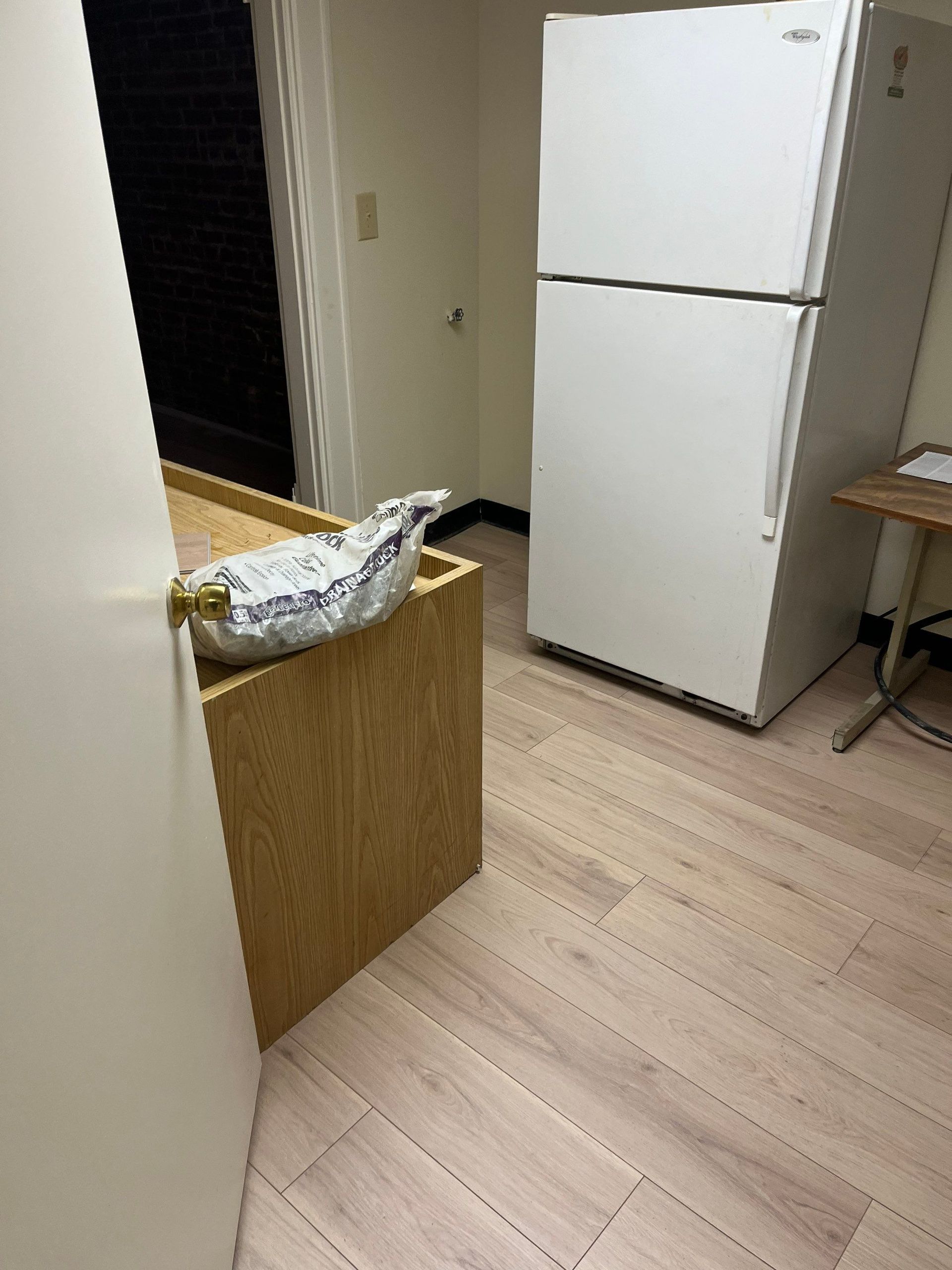 White refrigerator next to a light wood cabinet with a wrapped item, and a small table.