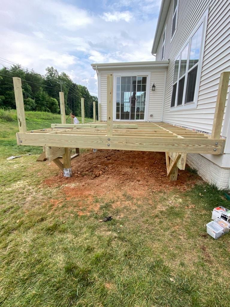 Partially built wooden deck next to a white house, on a grassy, dirt yard.