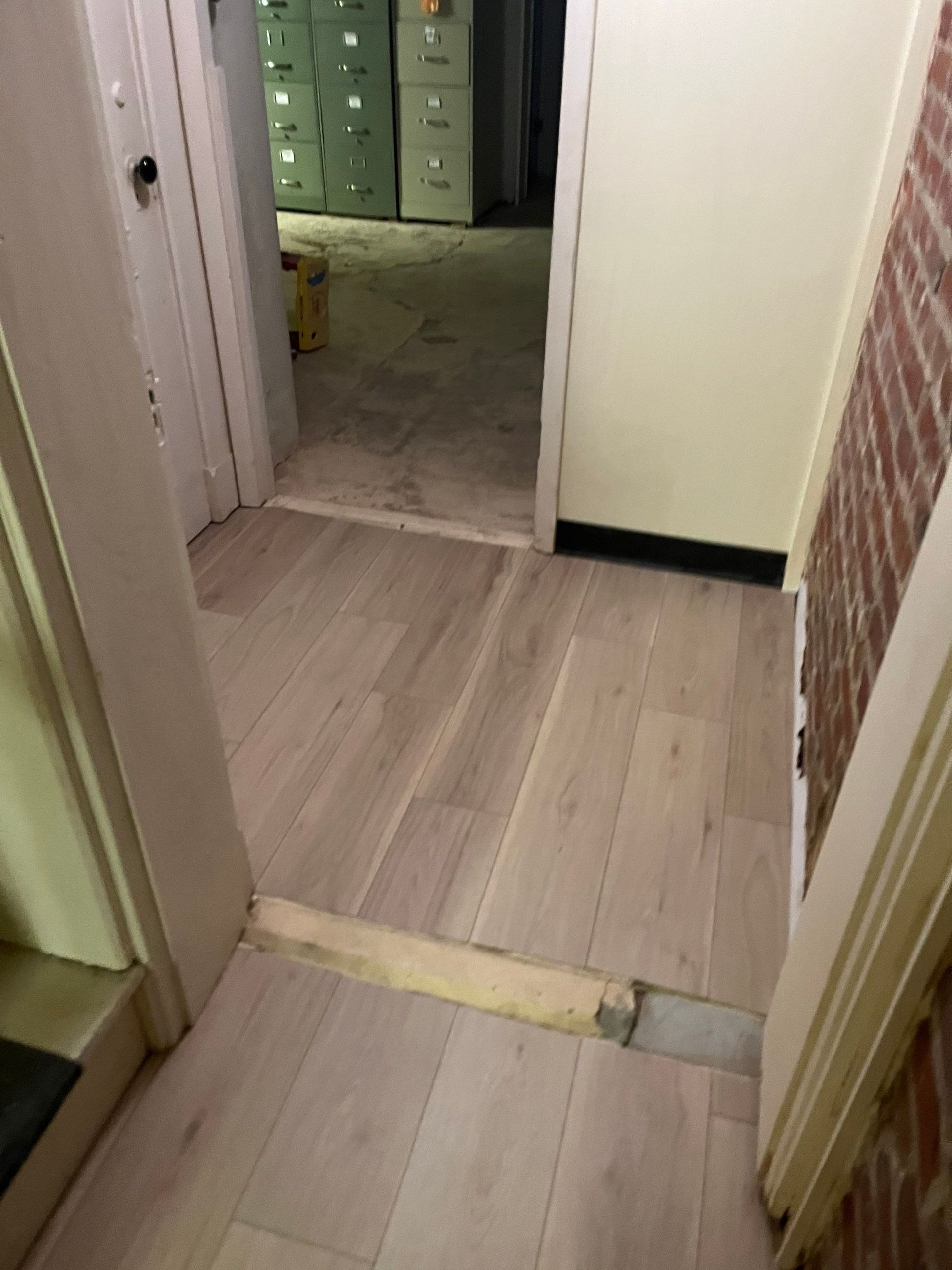 Hardwood floor leads to a doorway with brick wall and another room with filing cabinets in the background.