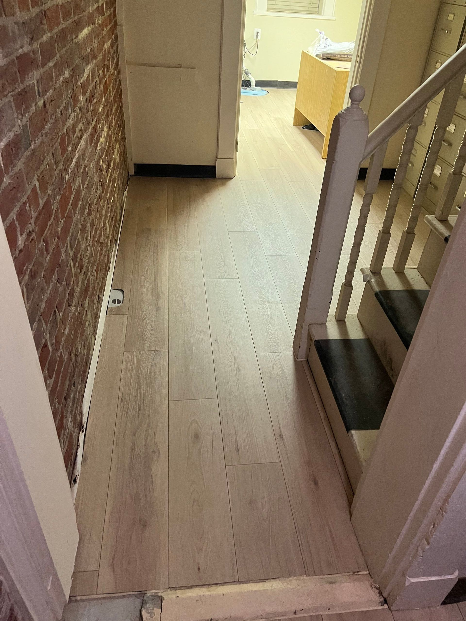 Hallway with light wood floor and brick wall, stairs on right, doorway ahead.