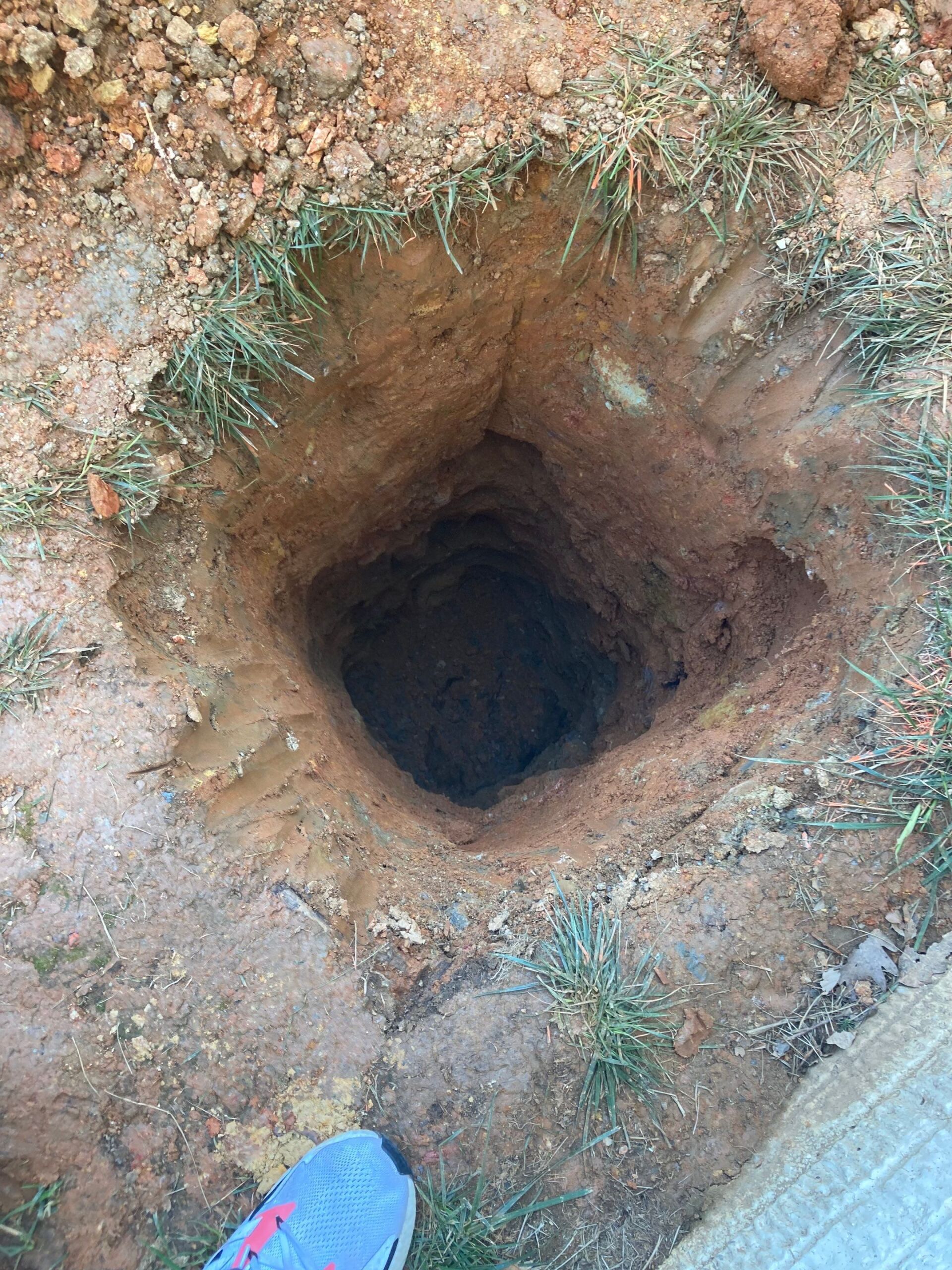 Square hole dug in the brown earth, surrounded by grass and dirt.