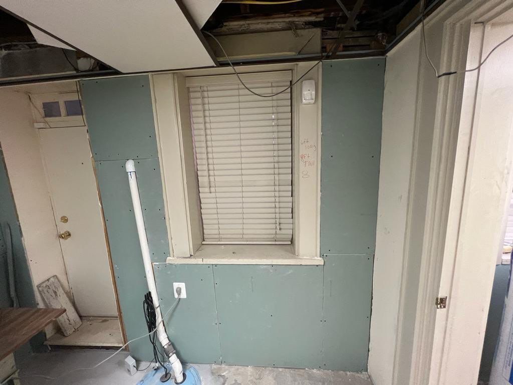 Basement under renovation with exposed ceiling, drywall, window with blinds, and doorway.