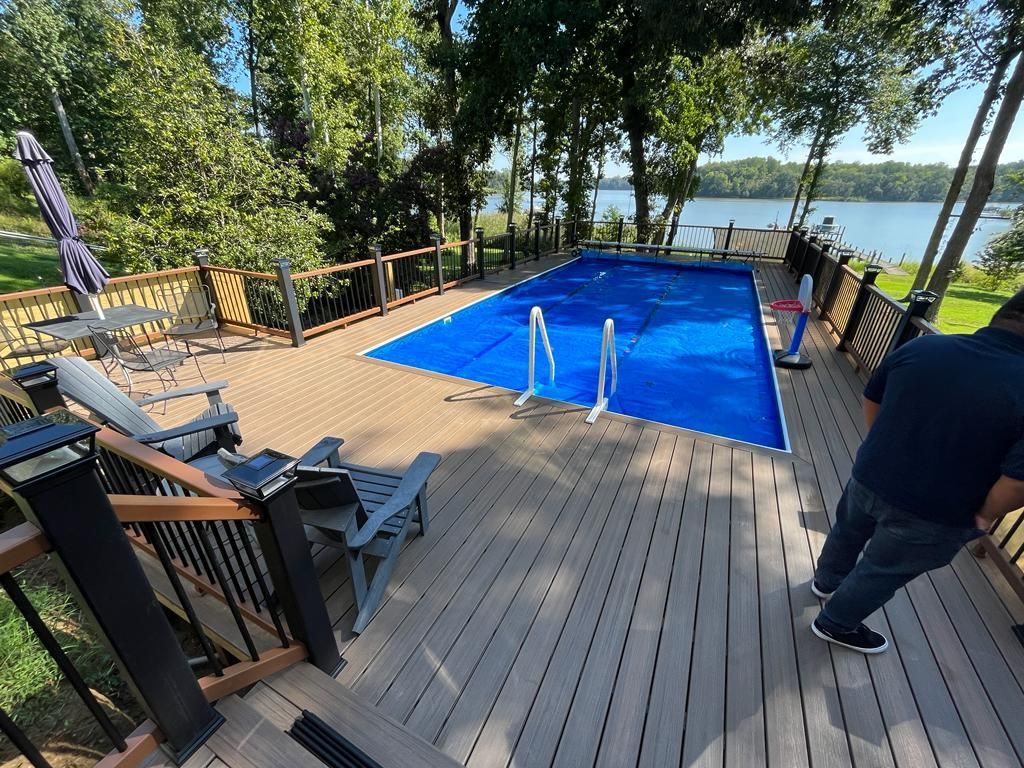 Deck with a blue-covered pool, overlooking a lake. A person moves on the deck. Trees and sky in the background.