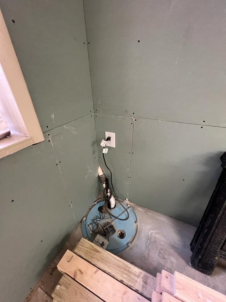 Sump pump in corner of a room, connected to electrical outlet. Light blue wall and blue container.