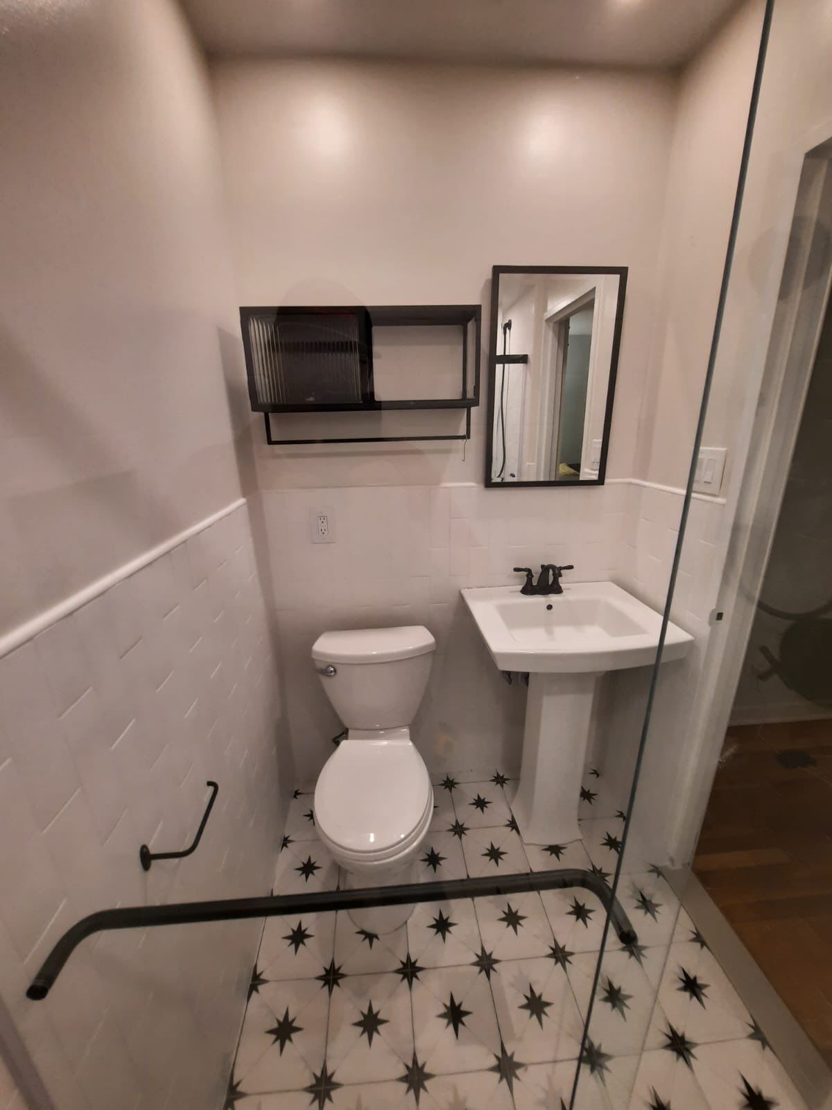 Small bathroom with white walls, black accents, and patterned tile floor; features toilet, sink, and shelf.