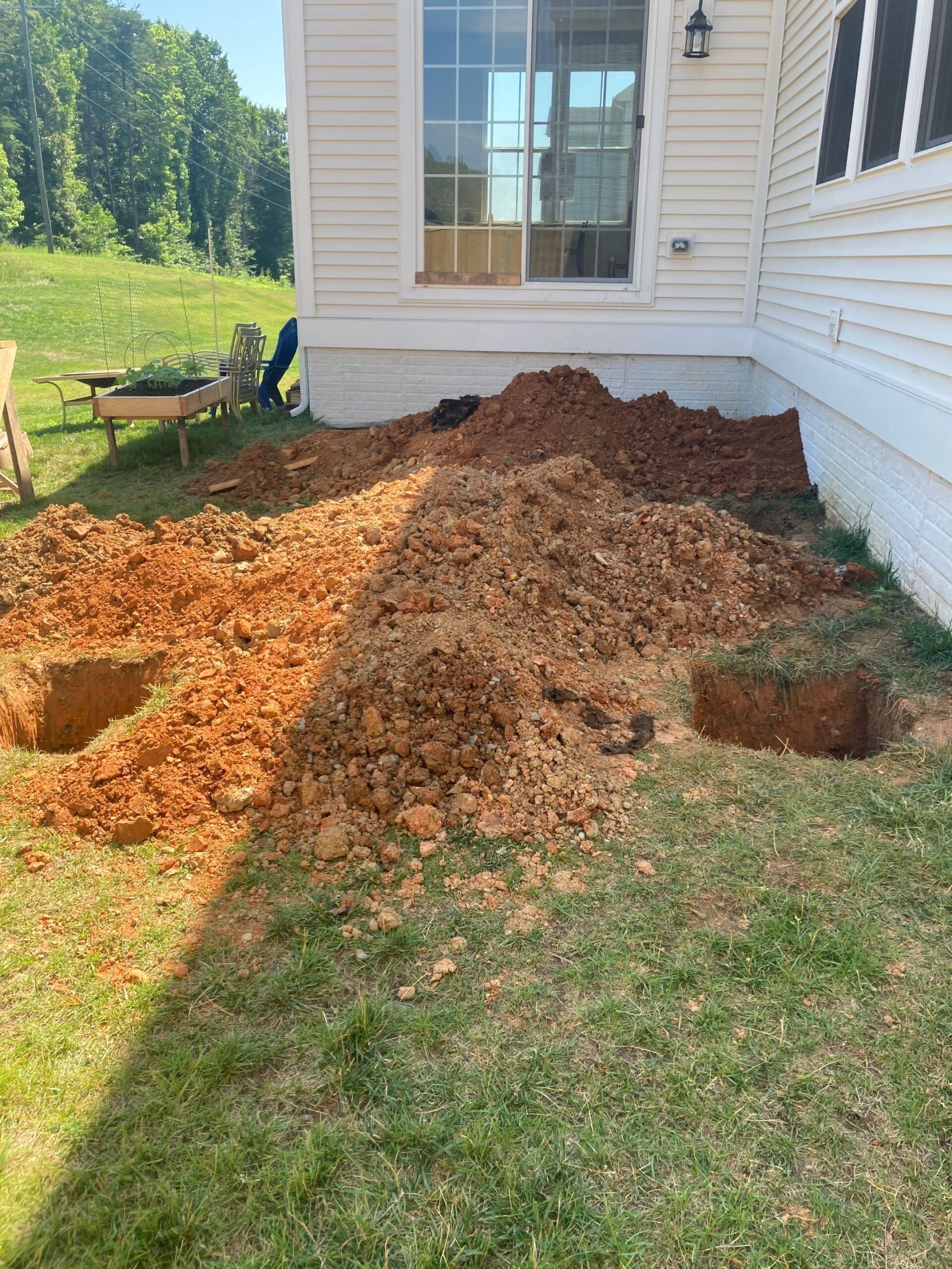 Three square holes dug in the ground near a house foundation, with piles of reddish-brown dirt.