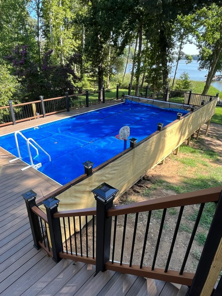 A backyard pool covered in blue with brown deck and fencing, trees and water in the background.