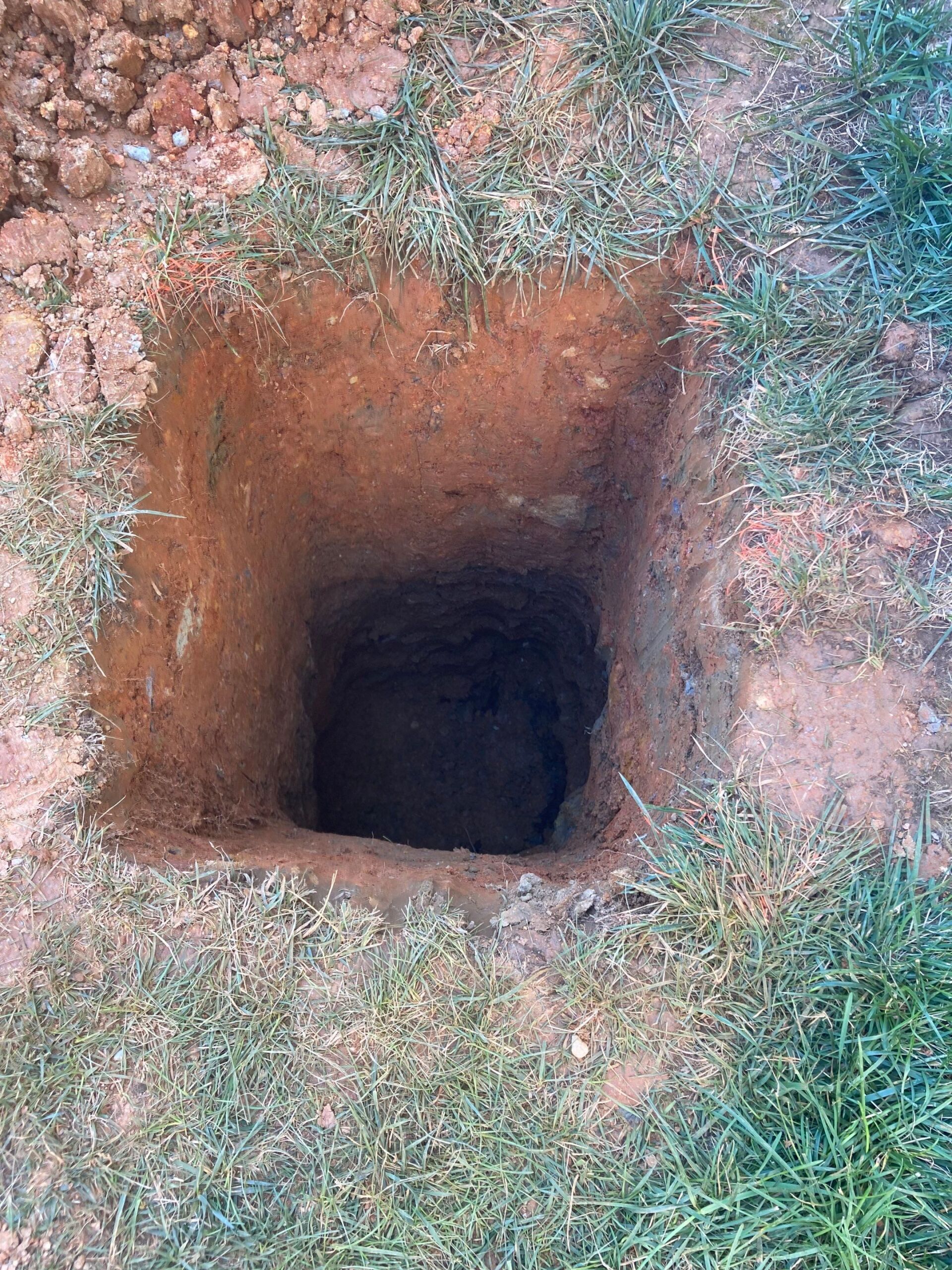 Square hole dug in the brown earth with surrounding green grass.