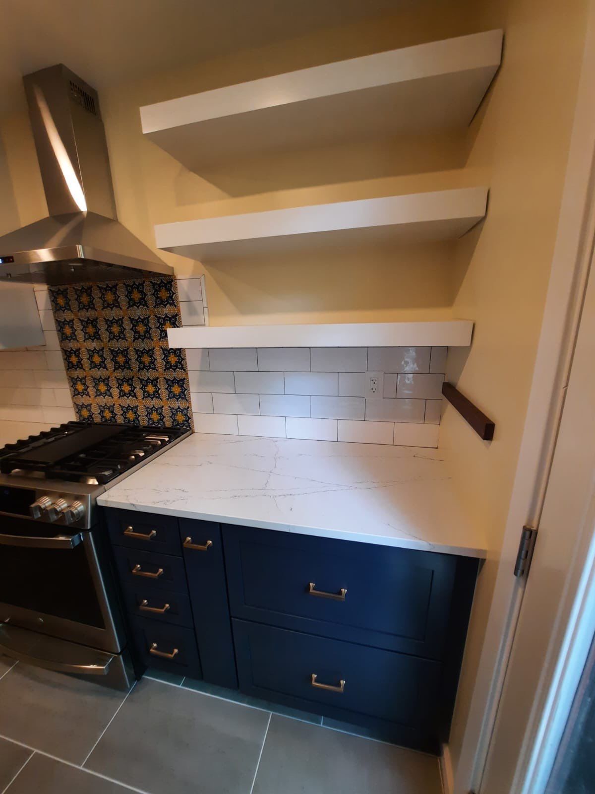 Kitchen renovation with navy blue cabinets, white countertops, floating shelves, and a tiled backsplash.
