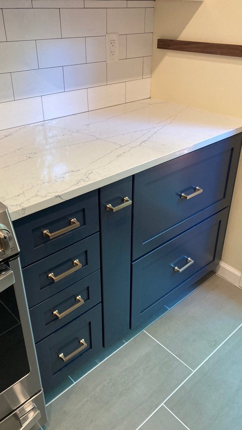 Navy blue kitchen cabinets with gold hardware and white countertop; gray tile floor.