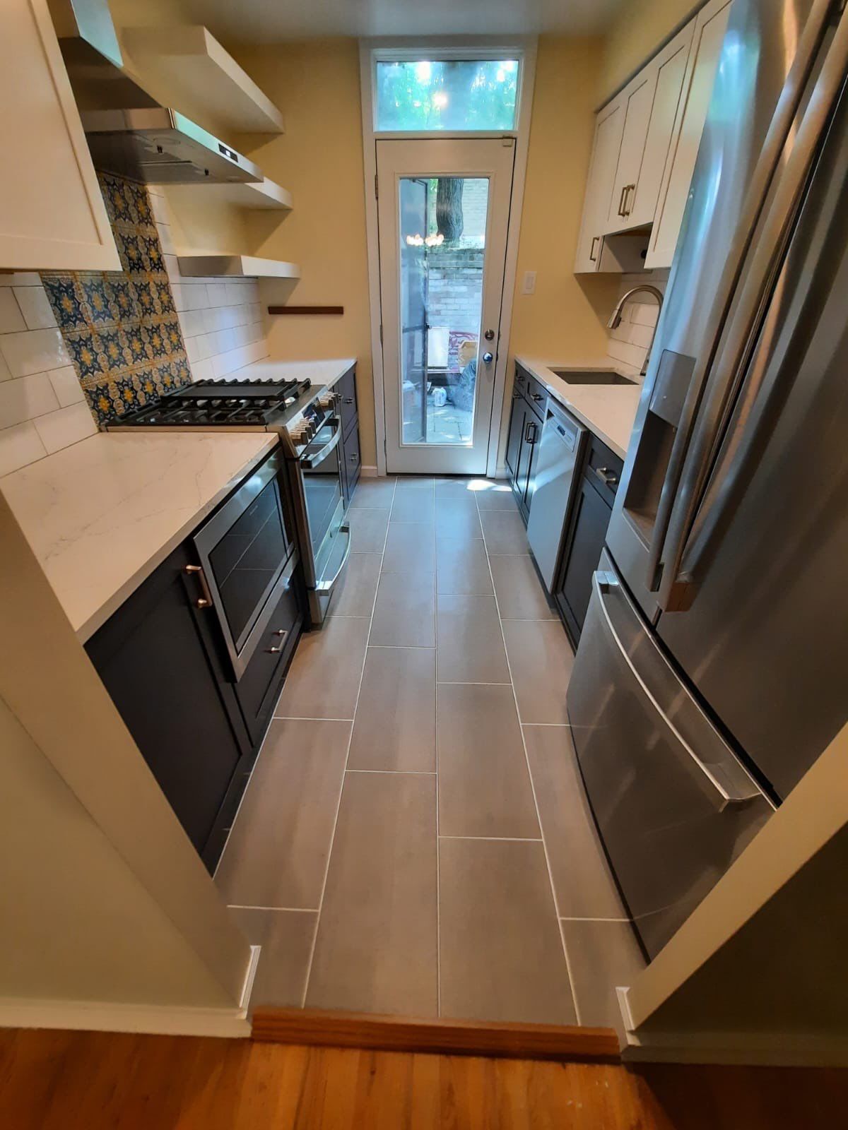Narrow kitchen with dark and light cabinets, gray tile floor, stainless steel appliances, and a door to the outside.