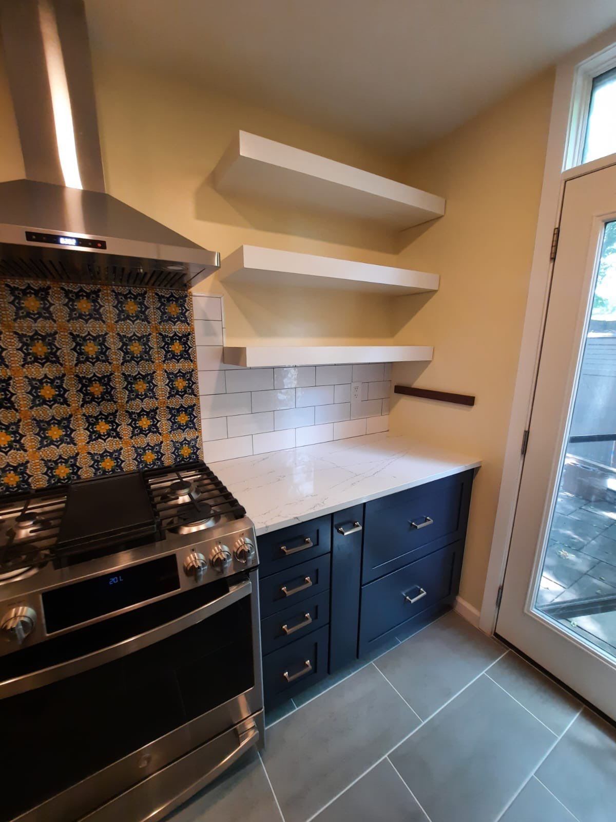Kitchen with blue cabinets, white countertops, floating shelves, and a stove with a patterned backsplash.