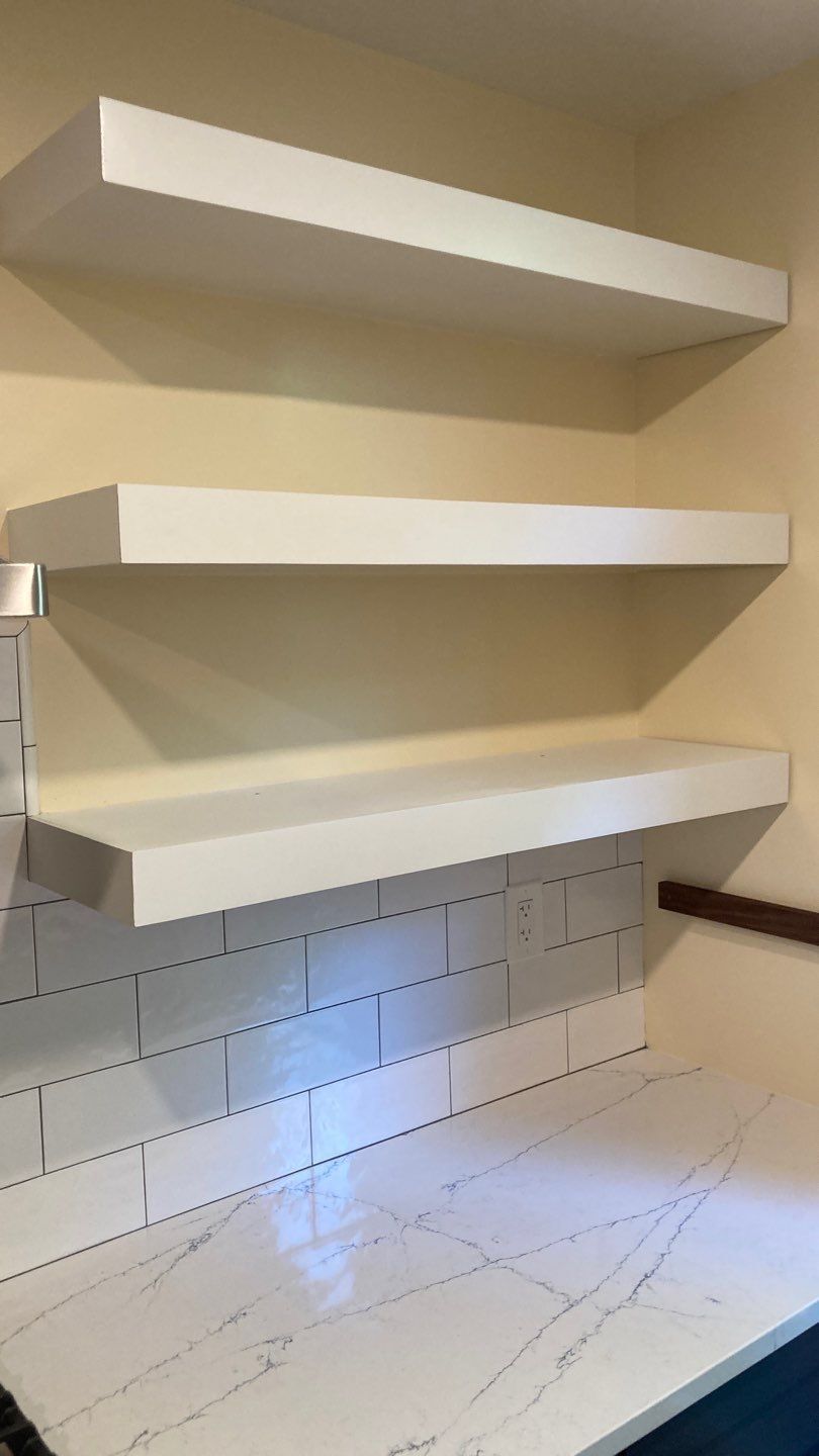 Three white floating shelves on a light beige wall above a countertop with white subway tile backsplash.