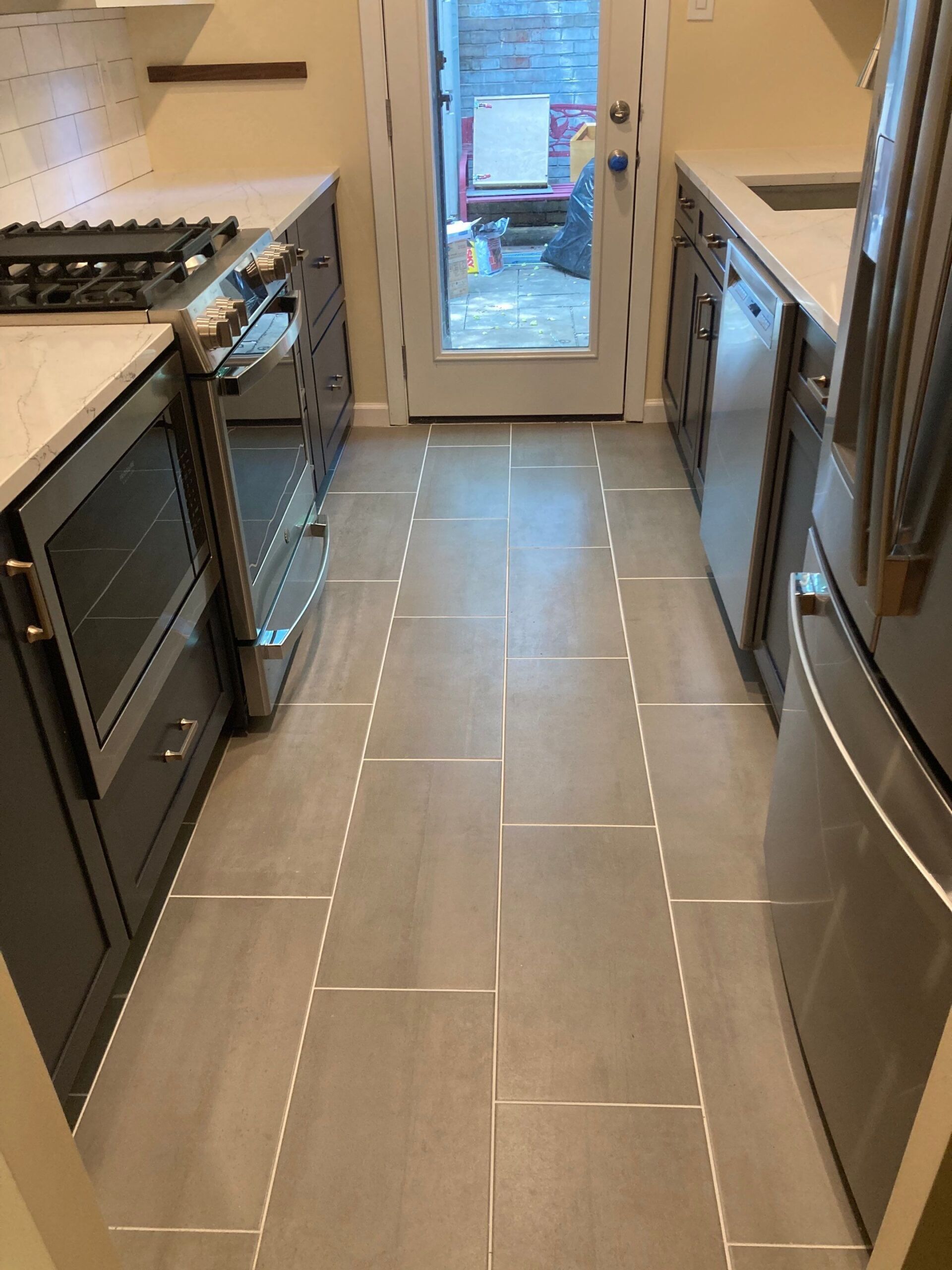 Narrow kitchen with gray tile floor, gray cabinets, stainless steel appliances, and door to outside.