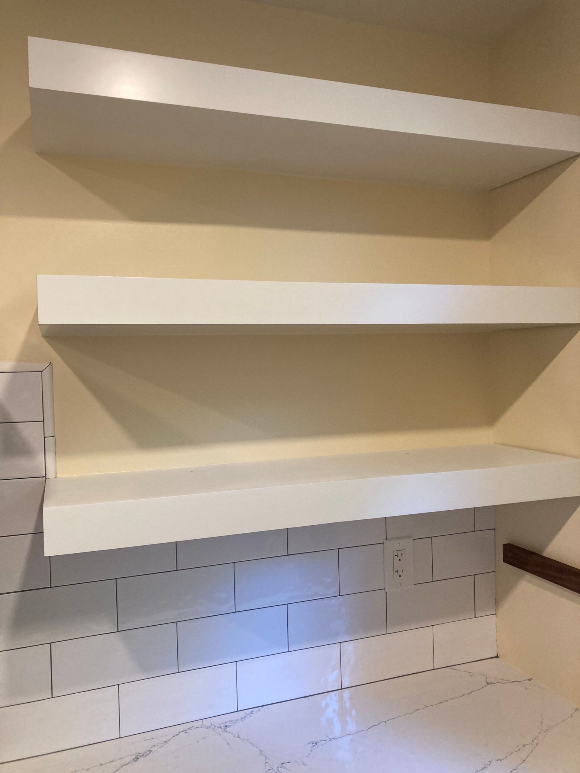 Three white shelves mounted on a beige wall. Below, a partially tiled area.