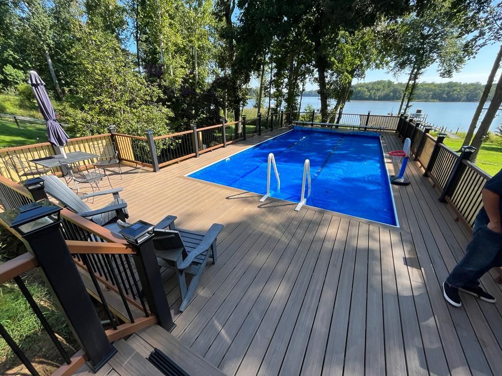 Wooden deck with pool, lake view, and trees. Blue pool cover, chairs, and railing.