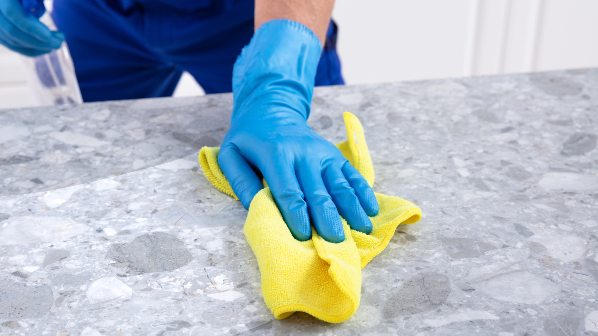 Person in a white shirt and blue apron smiling while cleaning on a sofa with spray bottle nearby