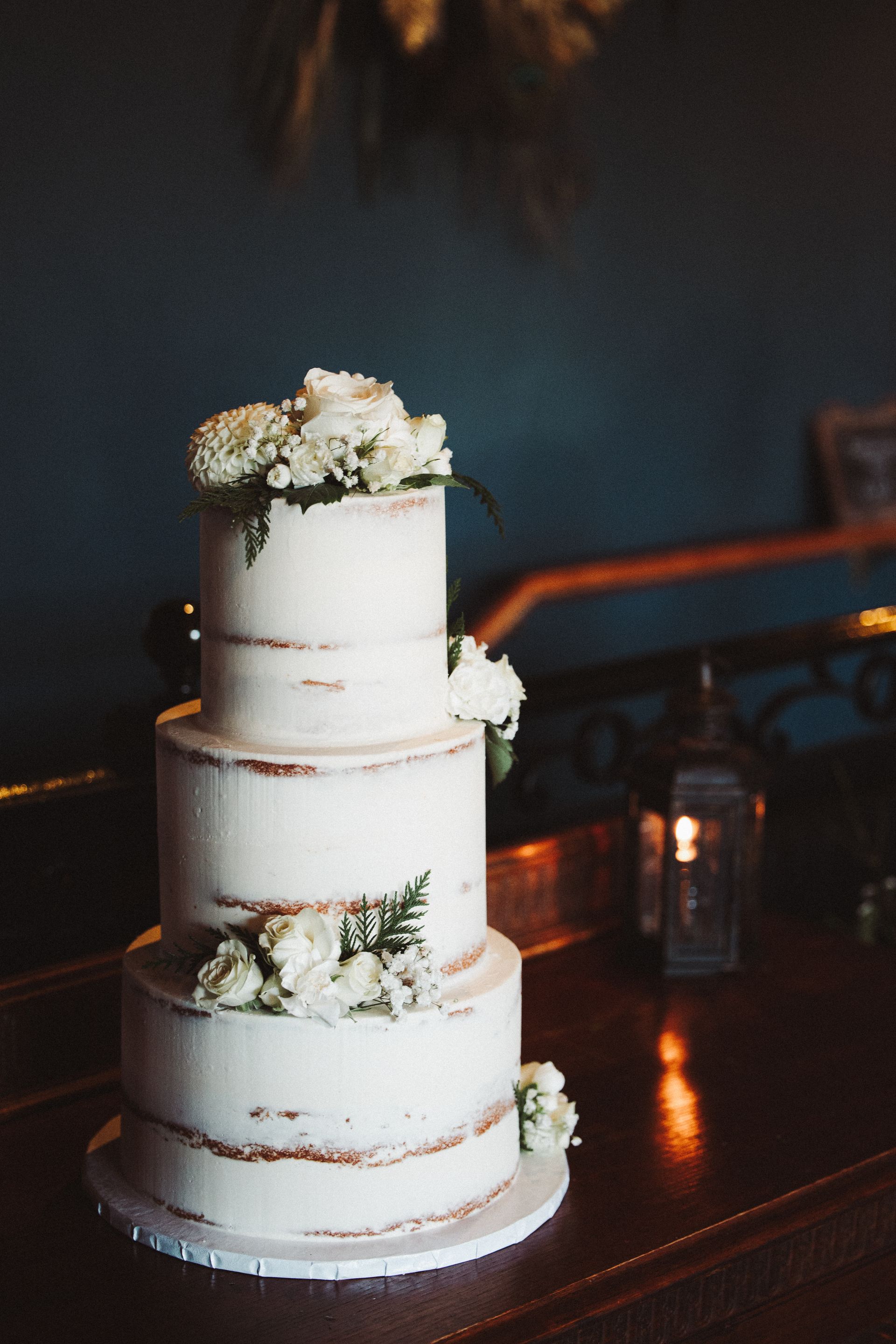 A three-tiered, white semi-naked wedding cake decorated with fresh white flowers and greenery on a wooden surface.