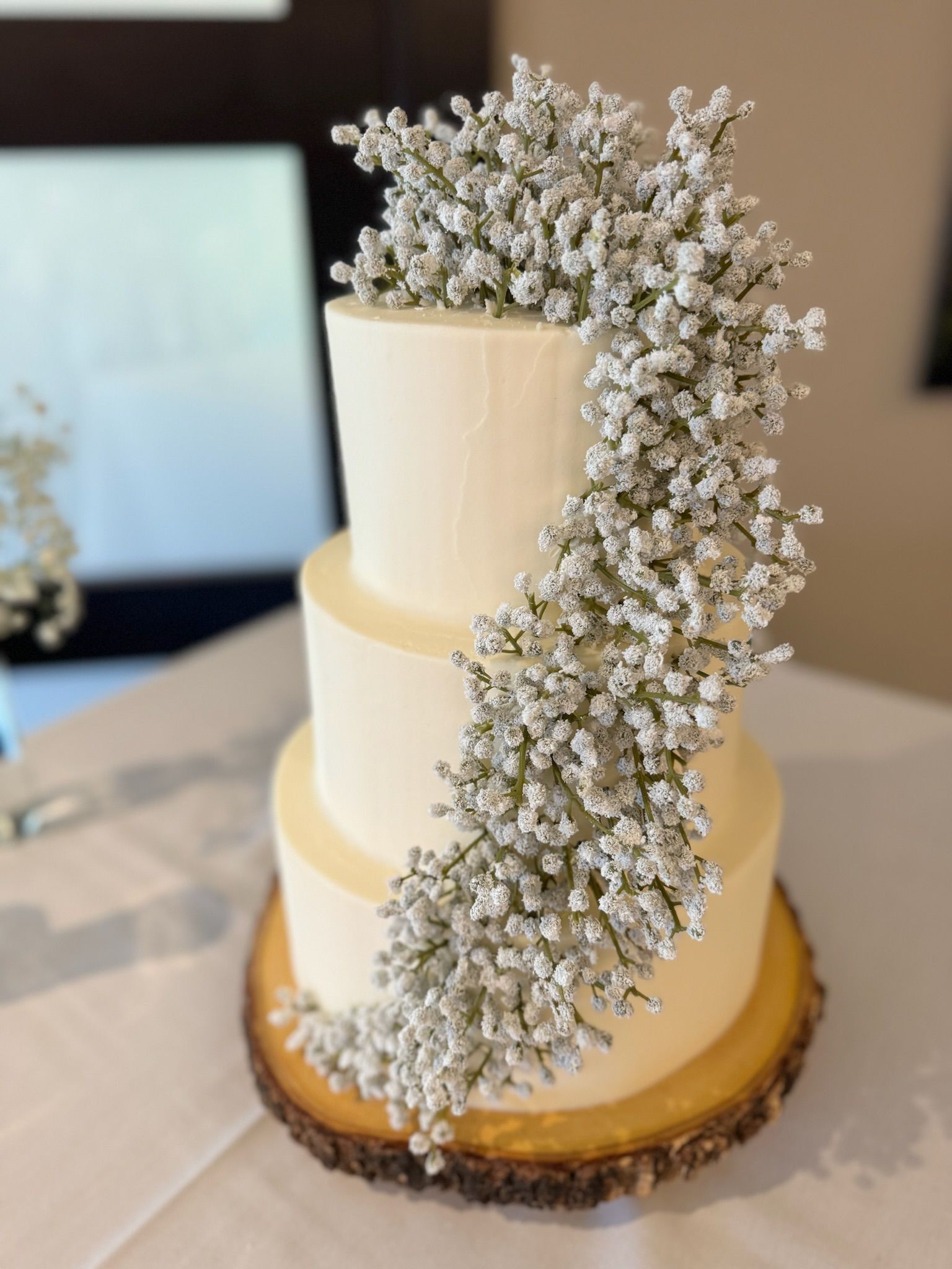 A white wedding cake with baby 's breath on top is sitting on a wooden slice on a table.