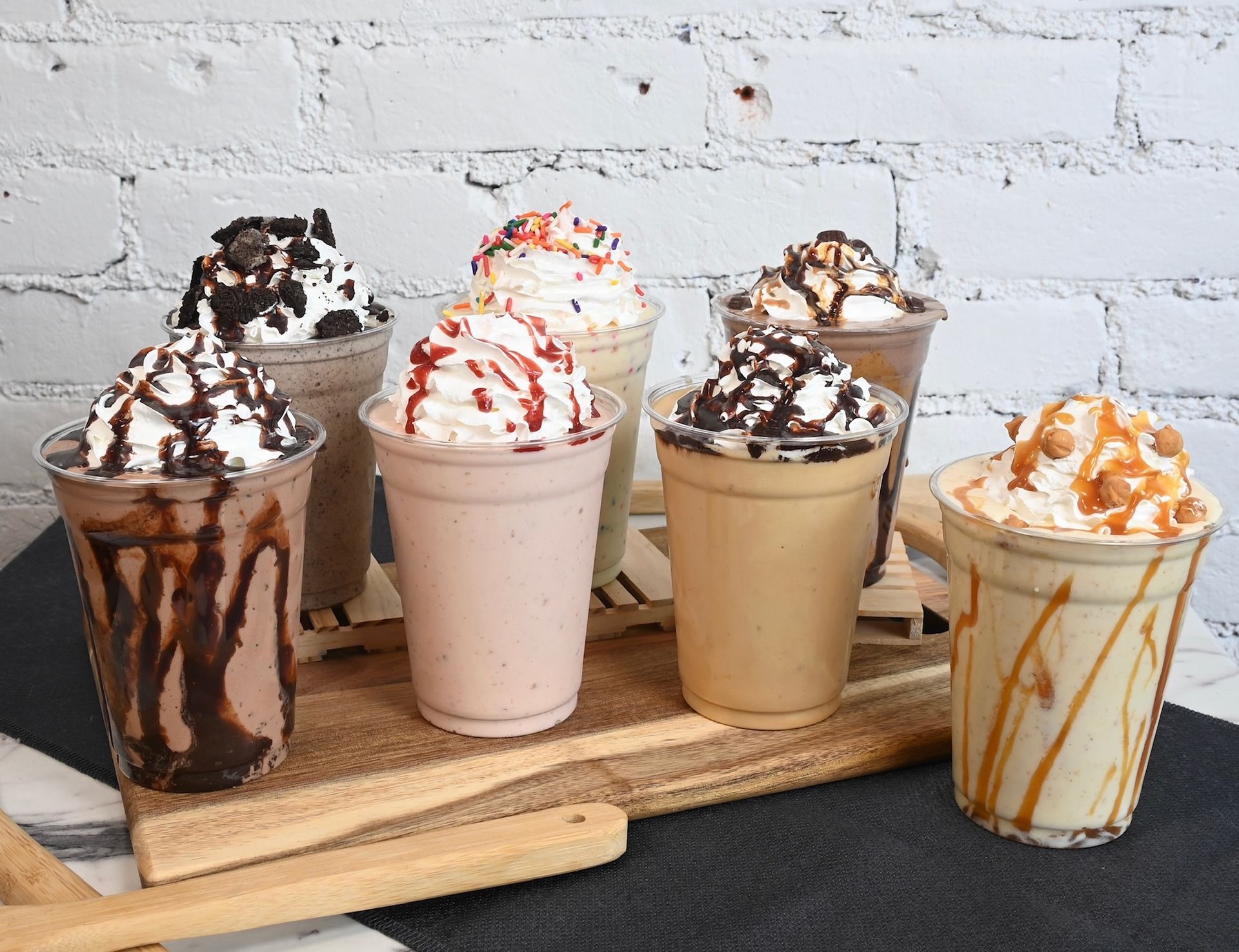 Five topped milkshakes in clear glasses on a wooden tray against a brick wall.