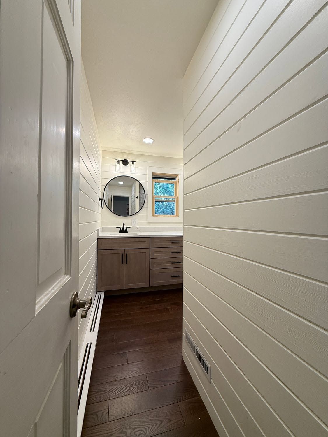 Narrow hallway to a bathroom vanity with round mirror, white paneled walls, and dark wood floor.