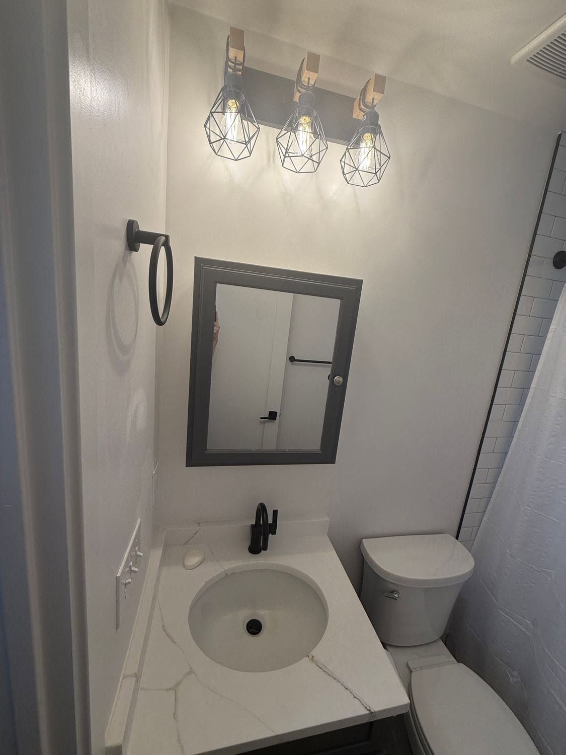 Small white bathroom with sink, mirror, toilet, shower curtain, and three-bulb light fixture.