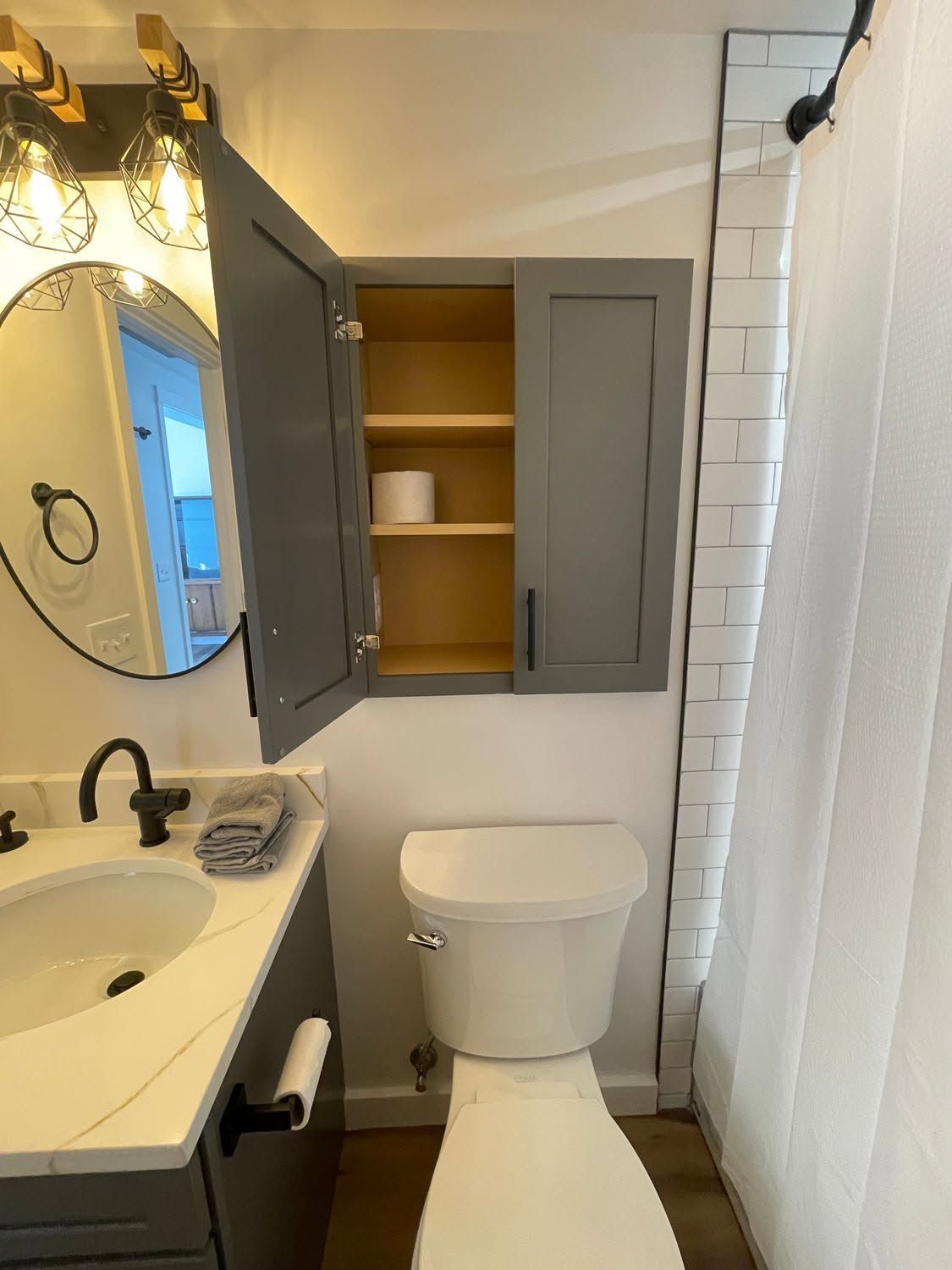 Small bathroom with open gray cabinet above toilet, sink vanity, mirror, and white shower curtain.