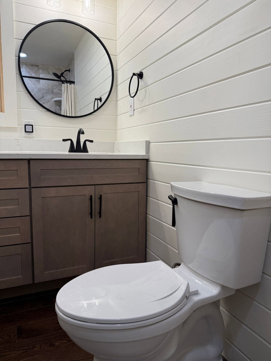 Small bathroom with white toilet, dark wood vanity, round mirror, and shiplap walls
