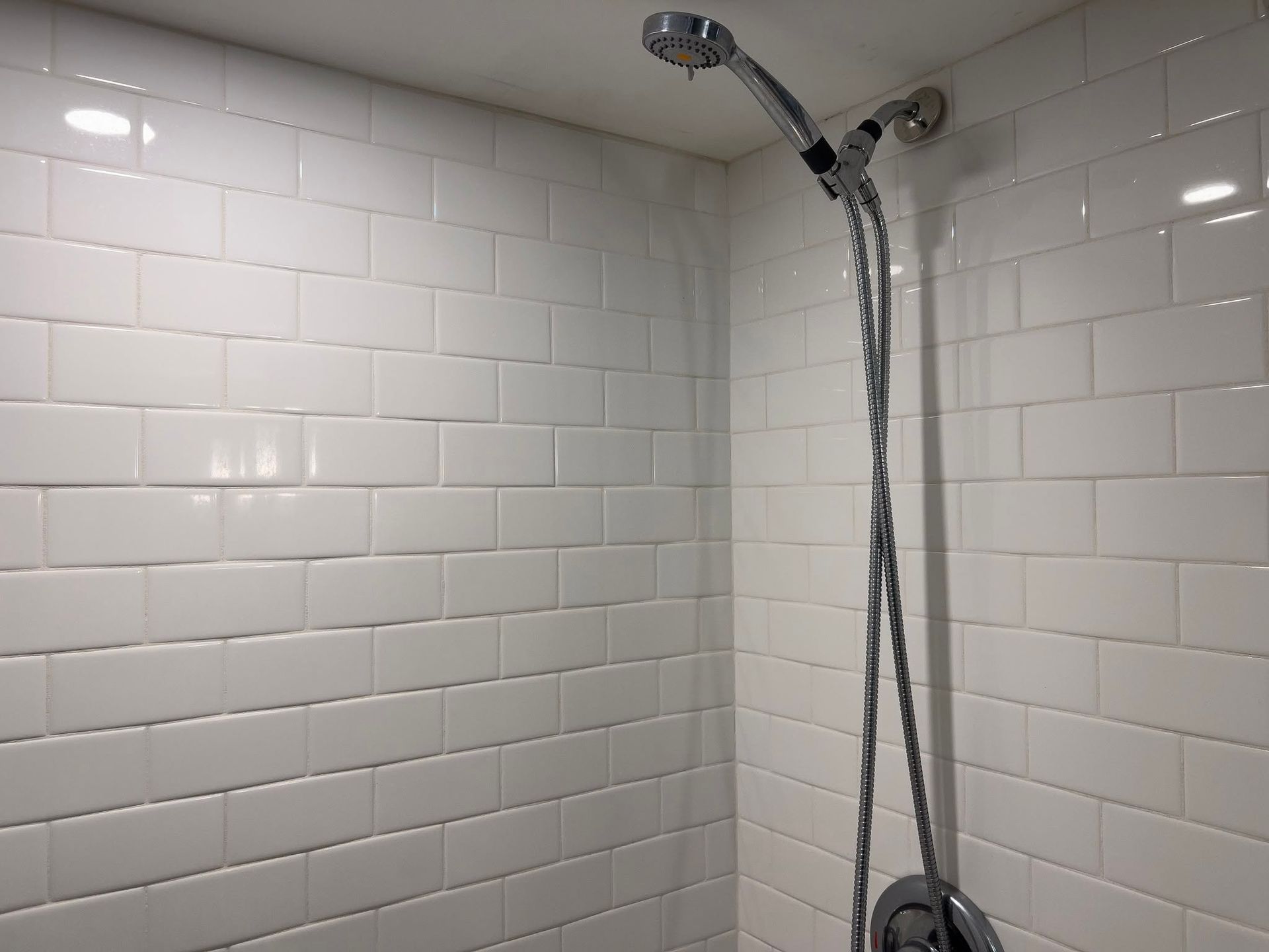 White-tiled shower corner with a chrome showerhead and handheld hose.