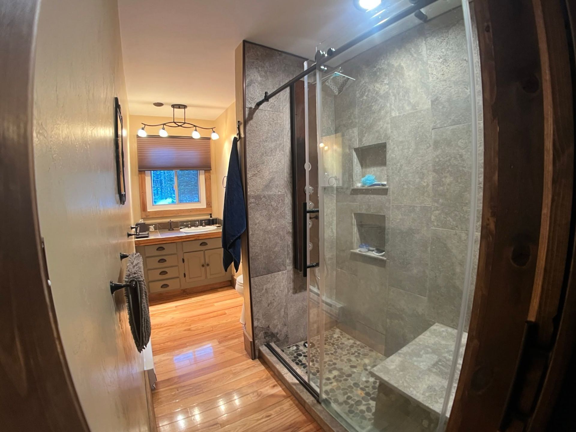 Narrow hallway bathroom with glass shower enclosure, wood vanity, and window at the far end