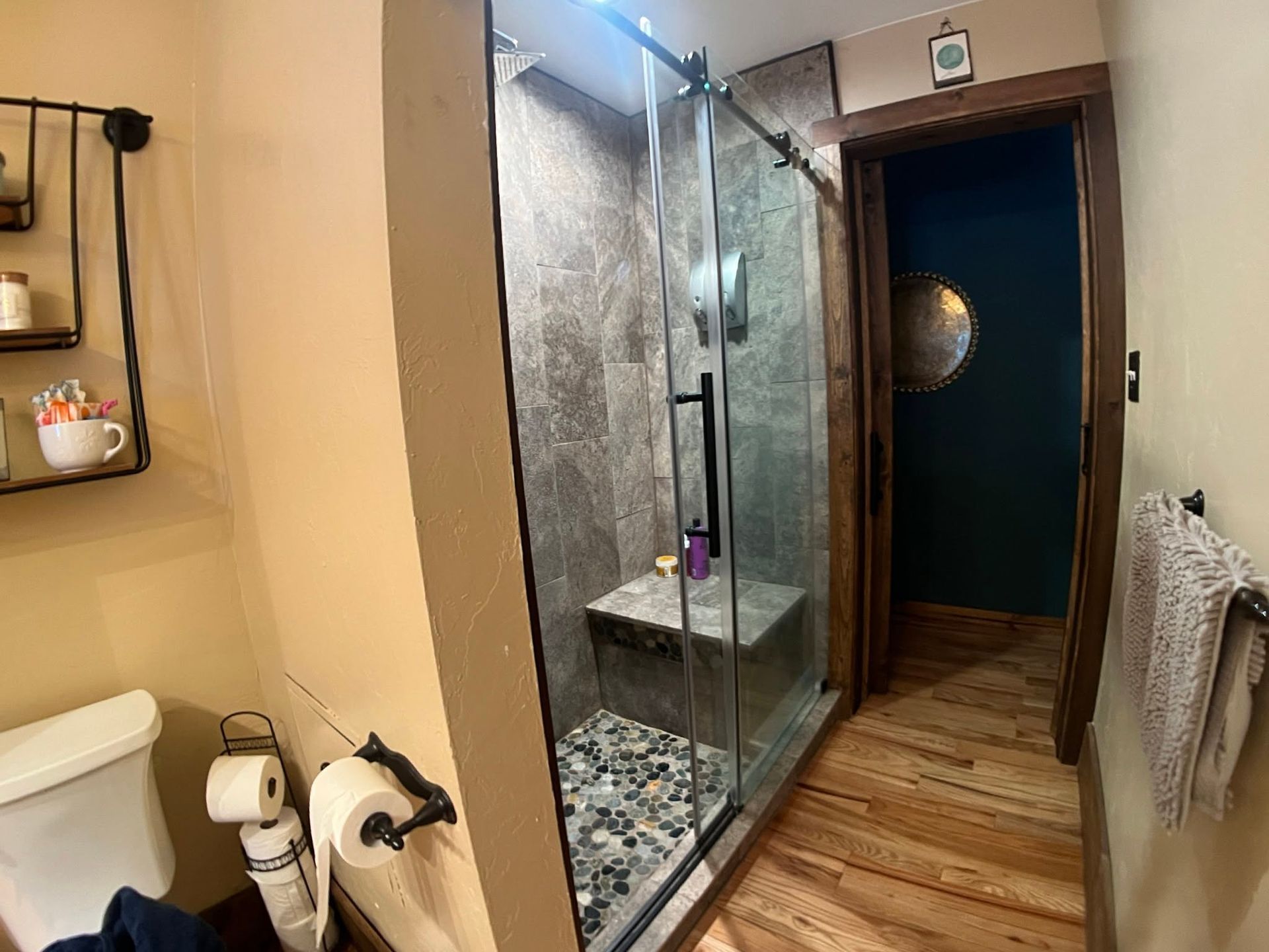 Bathroom with glass shower, white toilet, and doorway to a dark hallway with wood floors