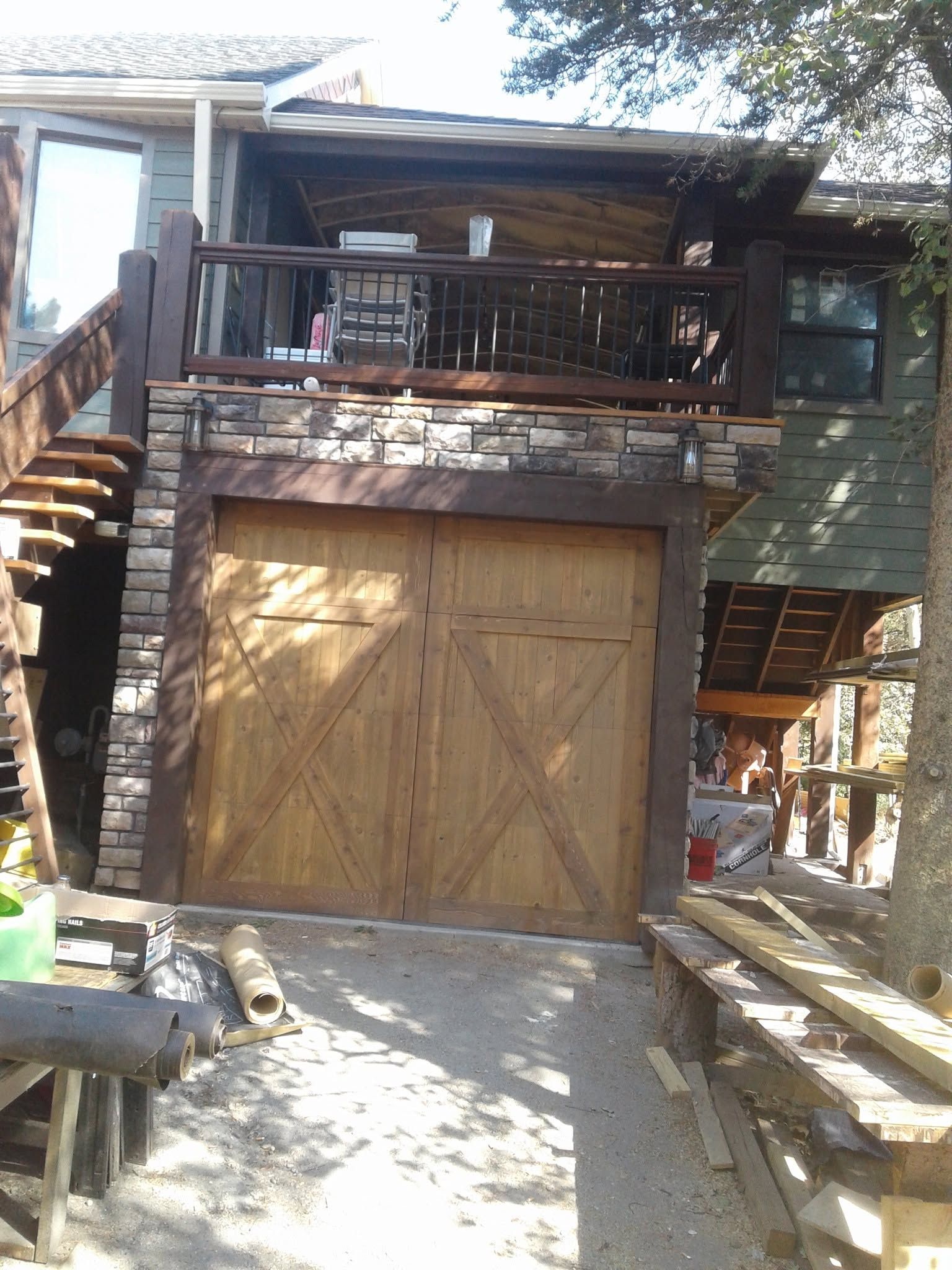 Wooden garage door beneath a stone porch and balcony on a rustic hillside home