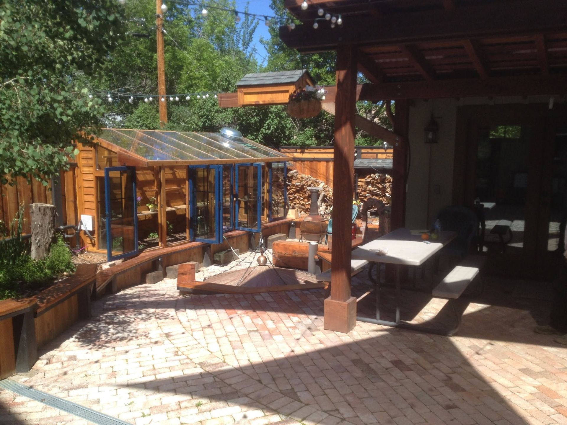 Sunlit patio with brick paving, picnic tables, and a wooden greenhouse beside a rustic building